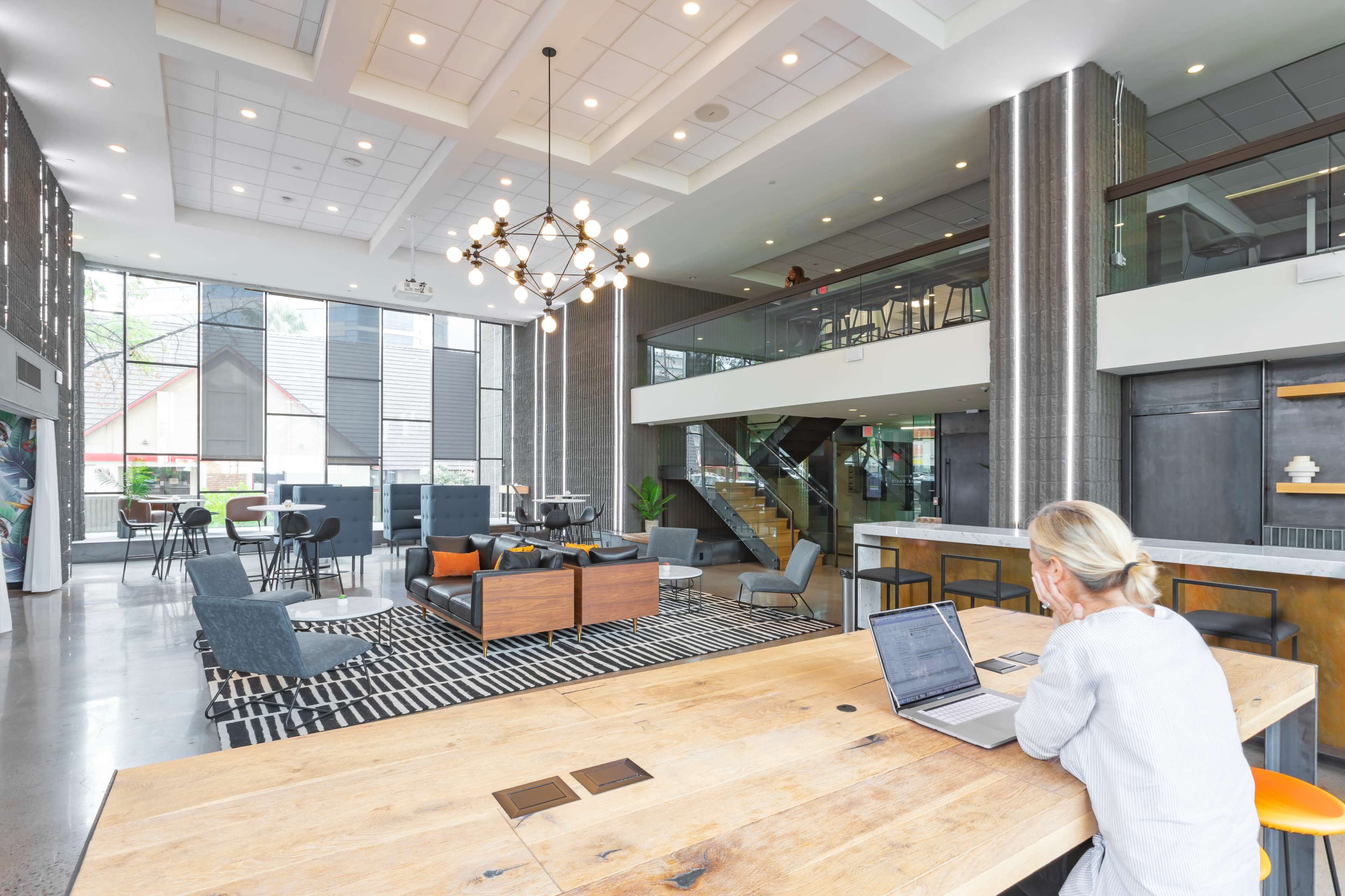 The image shows a modern open-concept lobby area with a combination of seating arrangements, a large staircase, and a person working on a laptop at a wooden table.