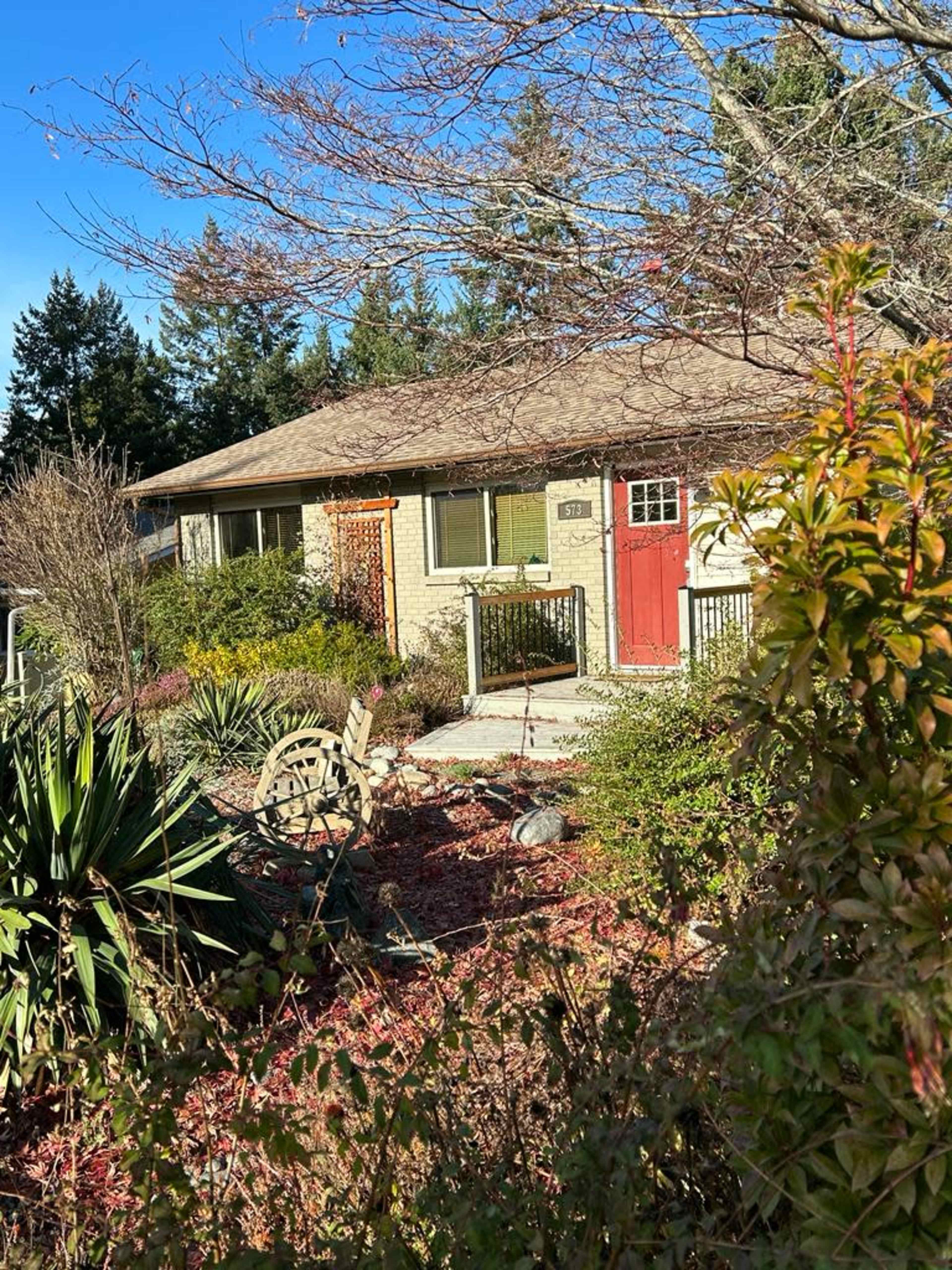A single-story house with a red door is surrounded by various shrubs and trees in a landscaped garden.