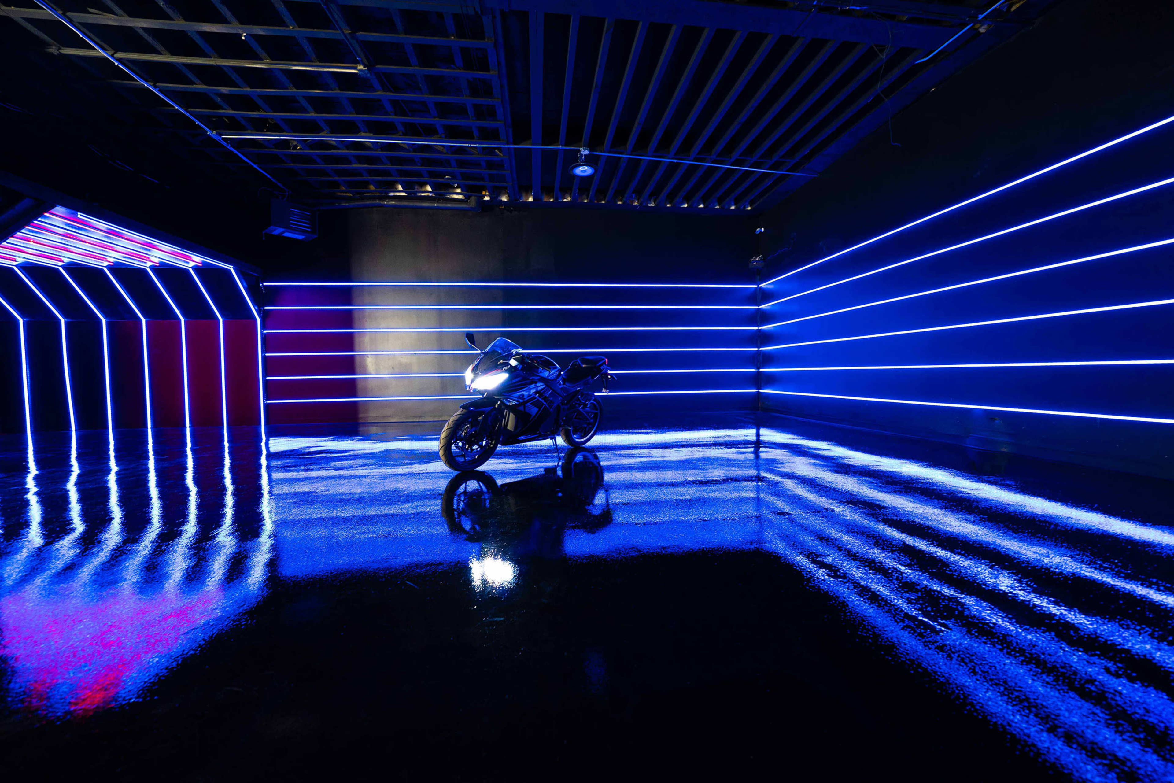 A motorcycle is positioned in a dark room illuminated by blue and purple neon lights, reflecting off the glossy floor.