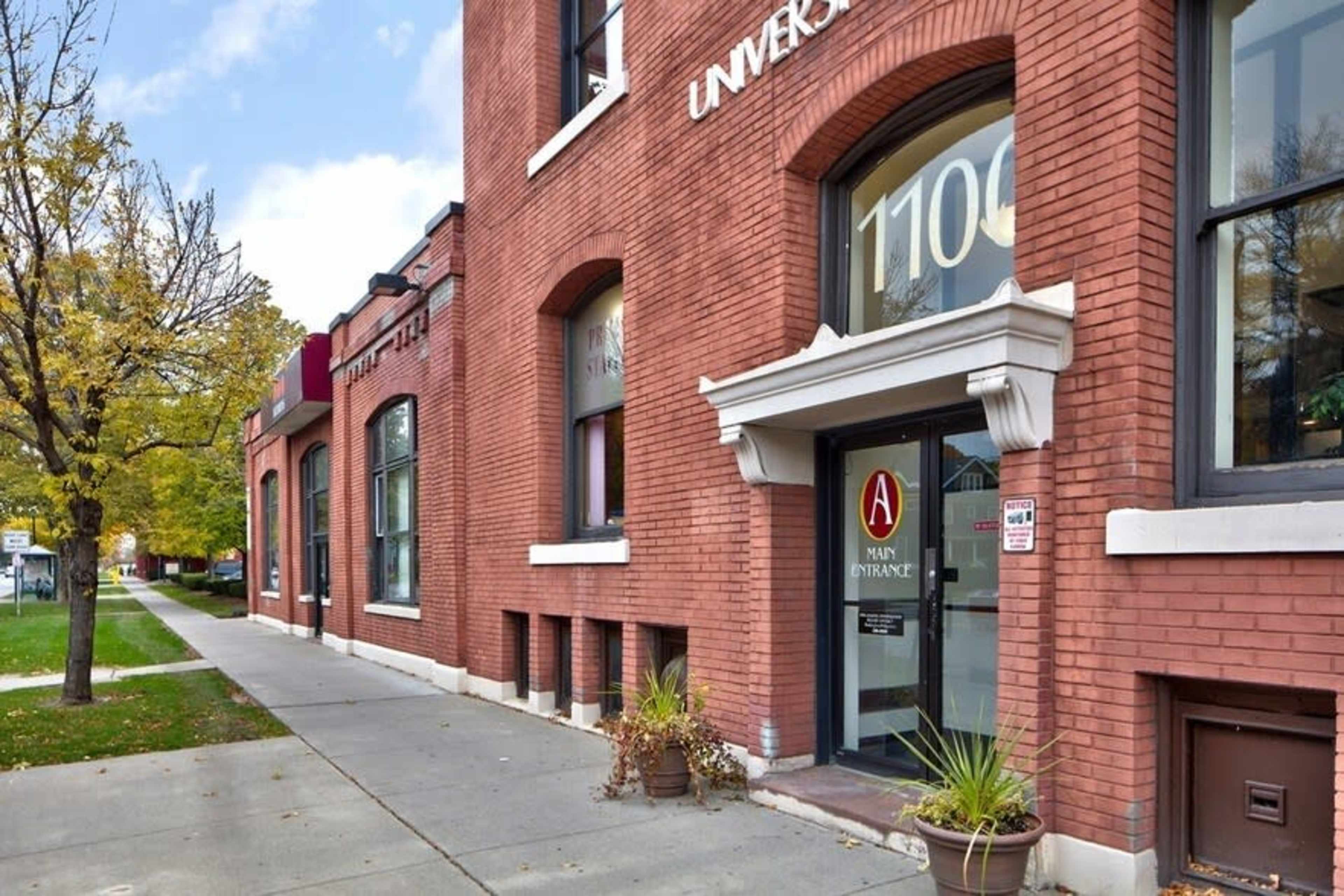 A brick building with large windows, featuring a main entrance marked by a sign and surrounded by planters, set along a sidewalk in an urban area.