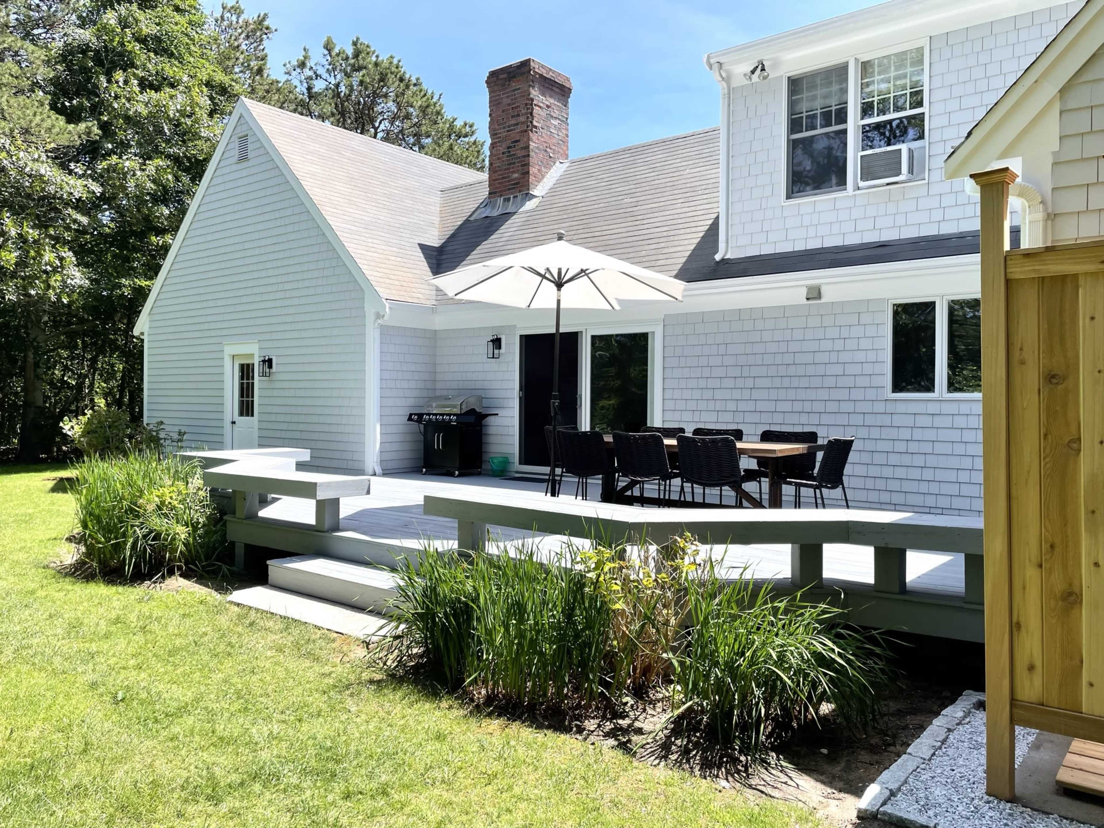 A modern house features a wooden deck with a dining area shaded by an umbrella, surrounded by a grassy lawn and trees.
