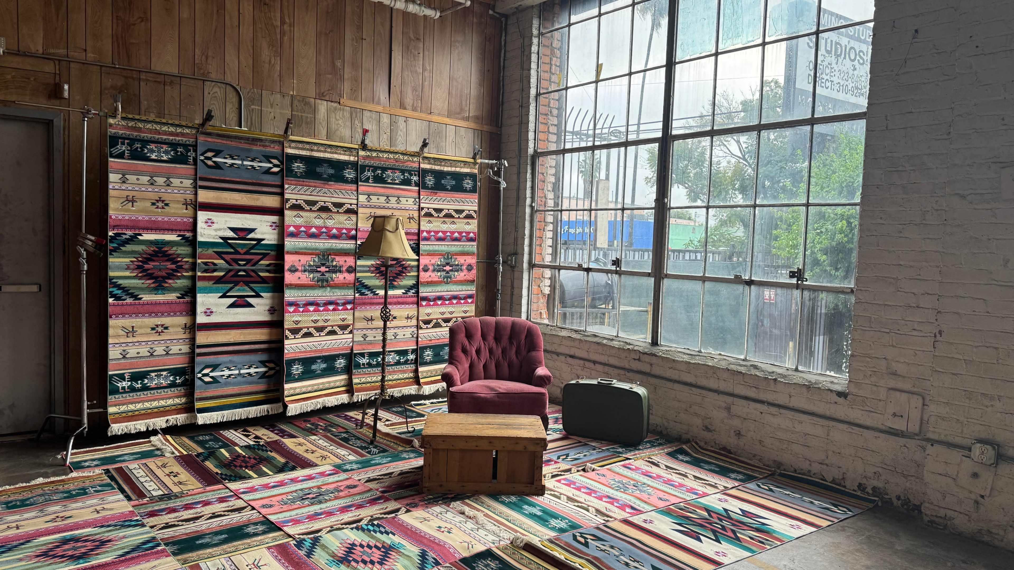The scene features a cozy, eclectic interior with a patterned area rug, a single maroon armchair, a wooden box, and decorative fabric panels against a large window with natural light filtering in.