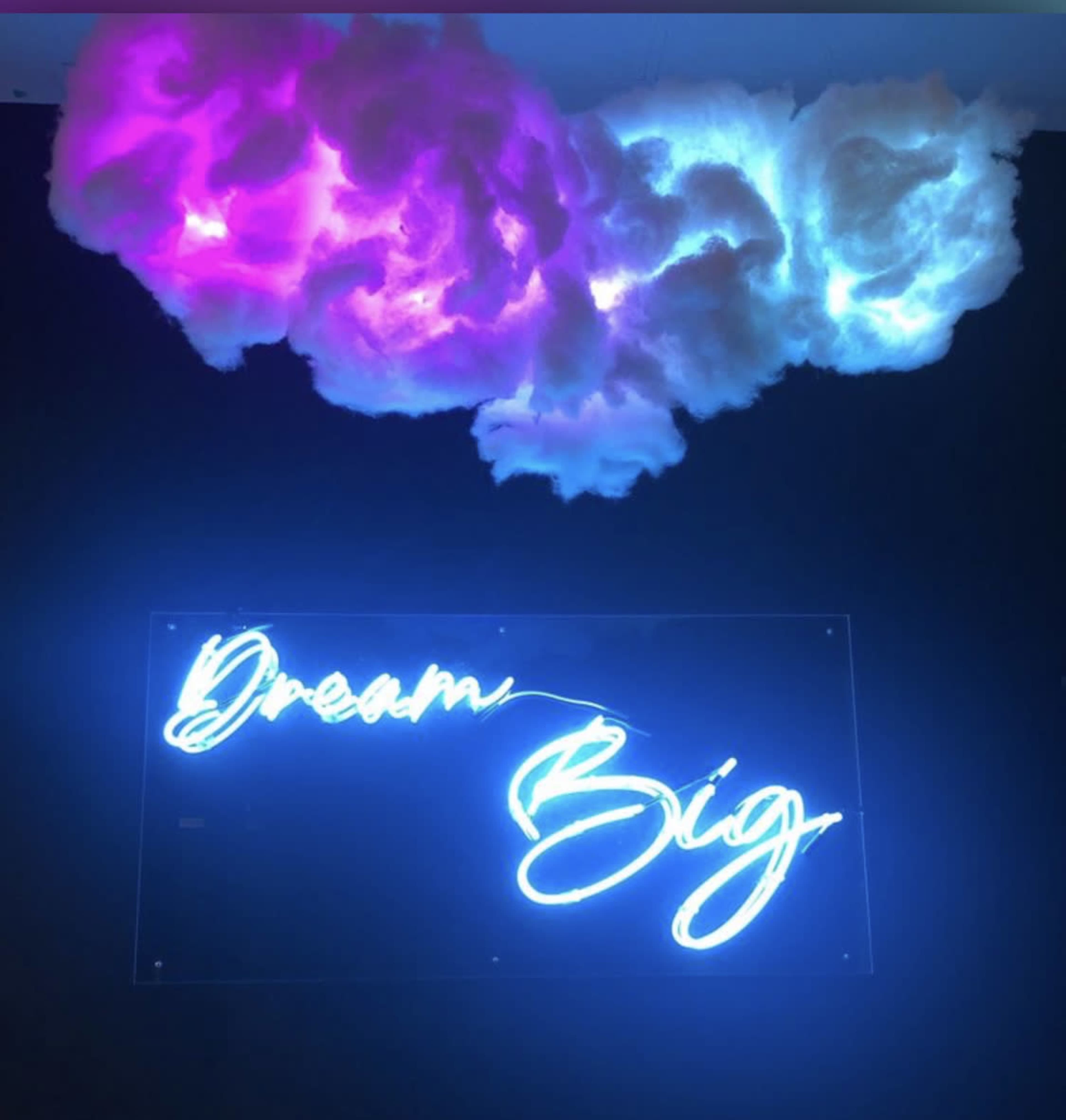 A neon sign that reads "Dream Big" beneath illuminated clouds in shades of pink and blue.