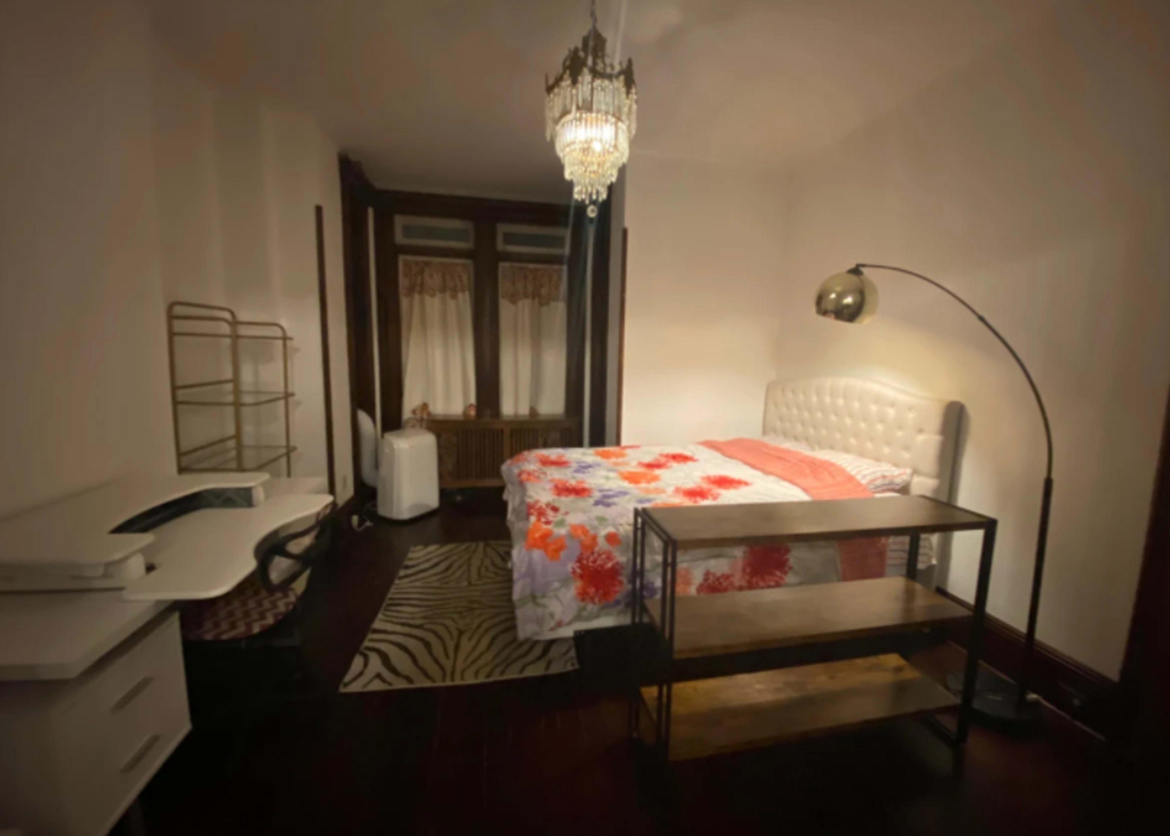 The image shows a bedroom featuring a bed with a floral comforter, a modern reading lamp, a small desk, and a glass top table.