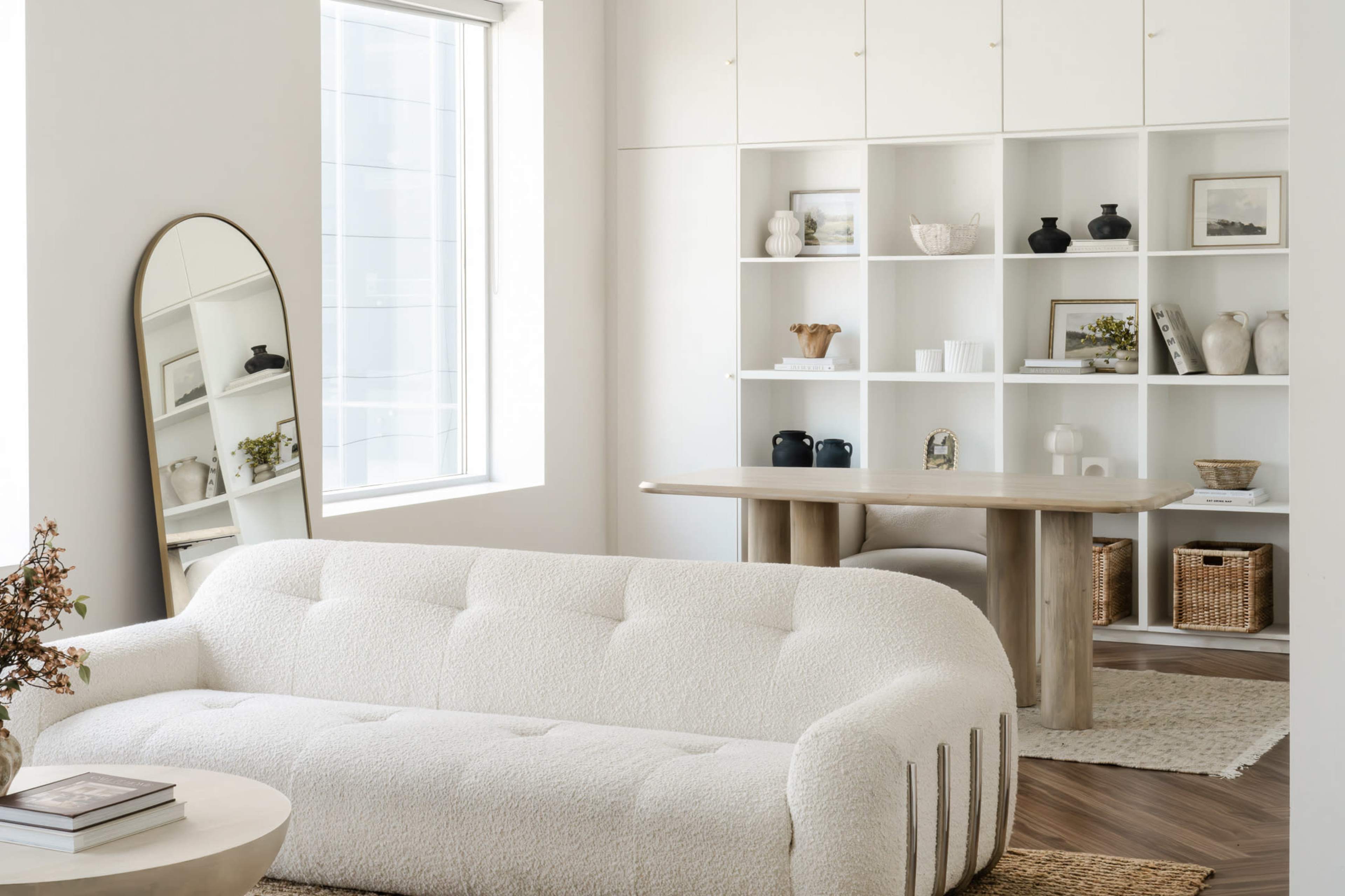The image shows a modern living room with a white curved couch, a circular coffee table, a dining area with a beige table, and shelves displaying various decorative items.