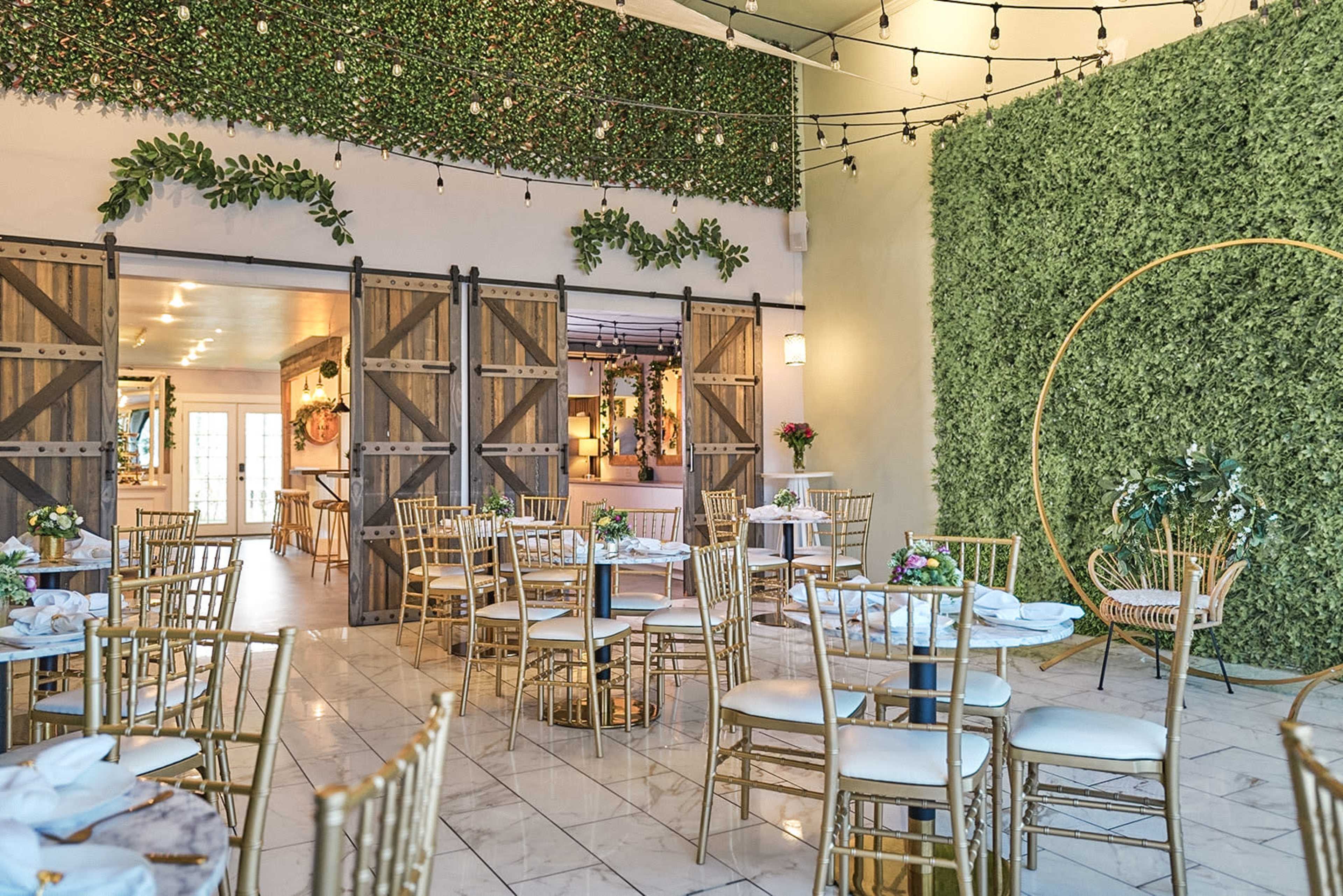 The image shows an elegantly decorated event space featuring gold chairs, round tables set with floral arrangements, and walls adorned with greenery and string lights.