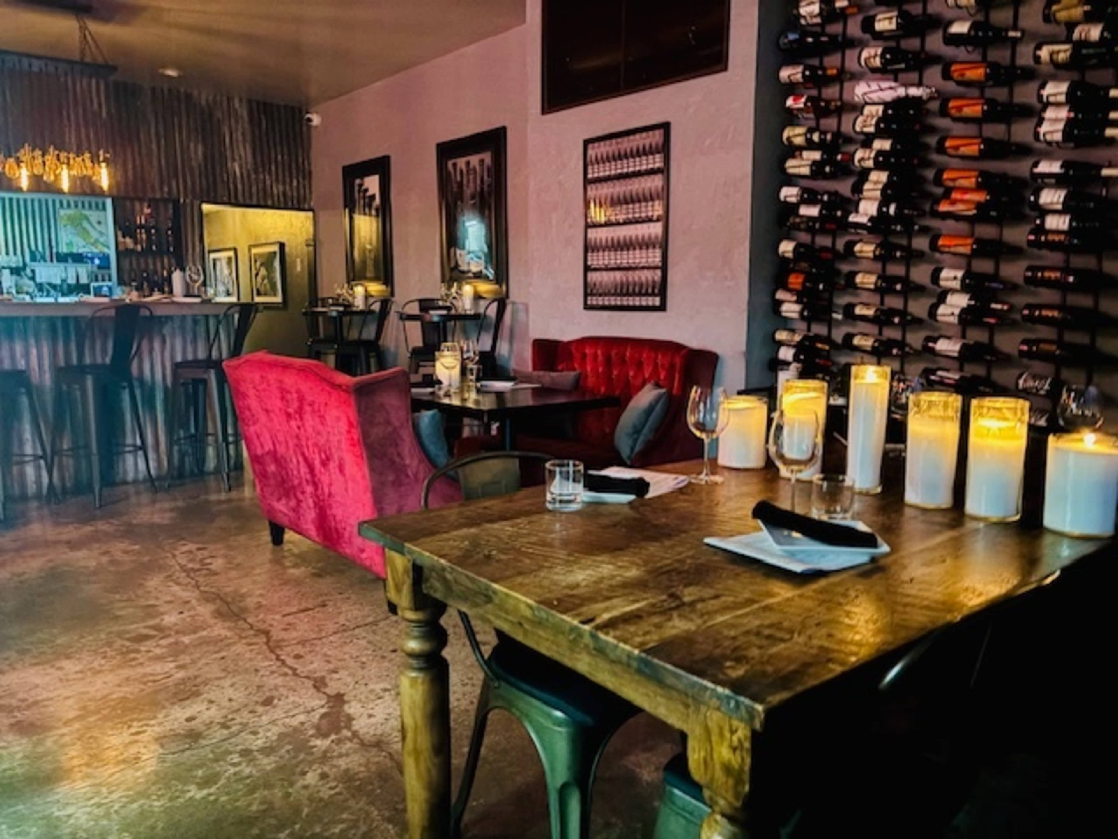 The image shows a dimly lit restaurant interior featuring wooden tables, red upholstered seating, and a wall displaying a wine rack.