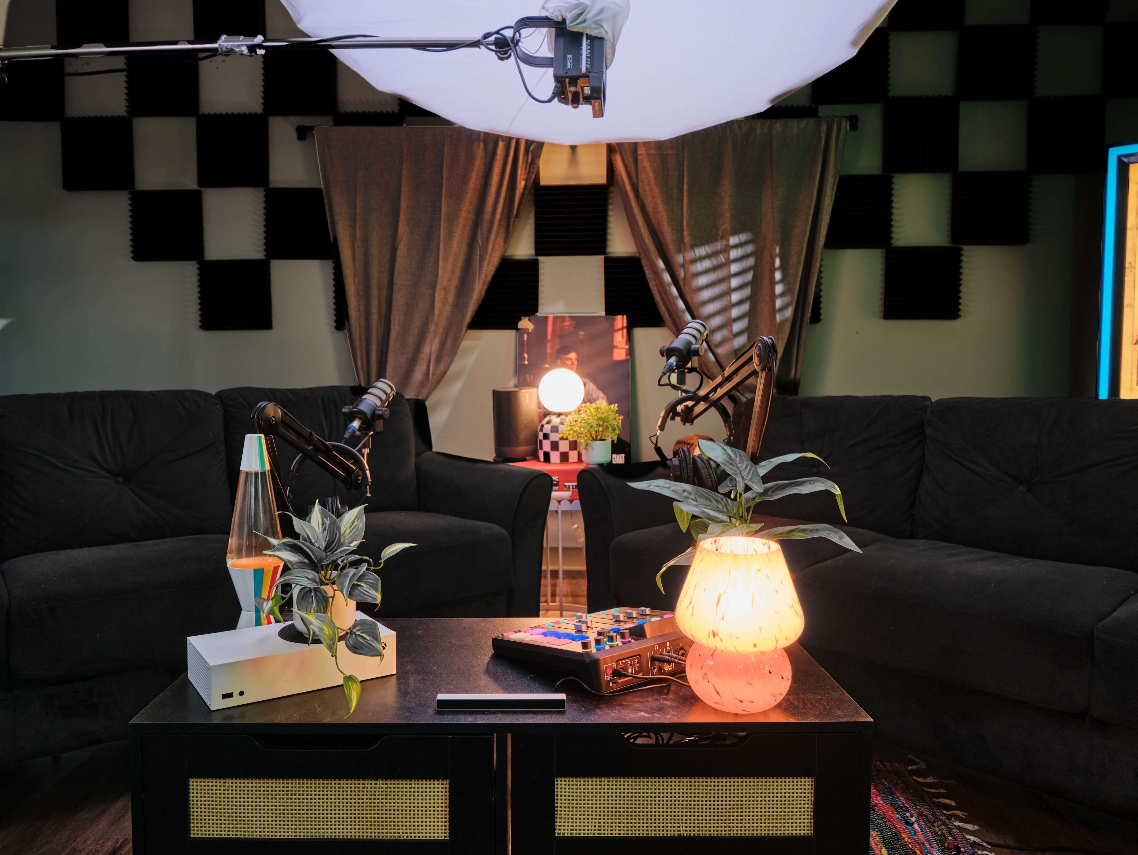 A cozy recording studio setup with two black couches facing a table, equipped with audio equipment, a lamp, and potted plants, under soft lighting and acoustic paneling on the walls.