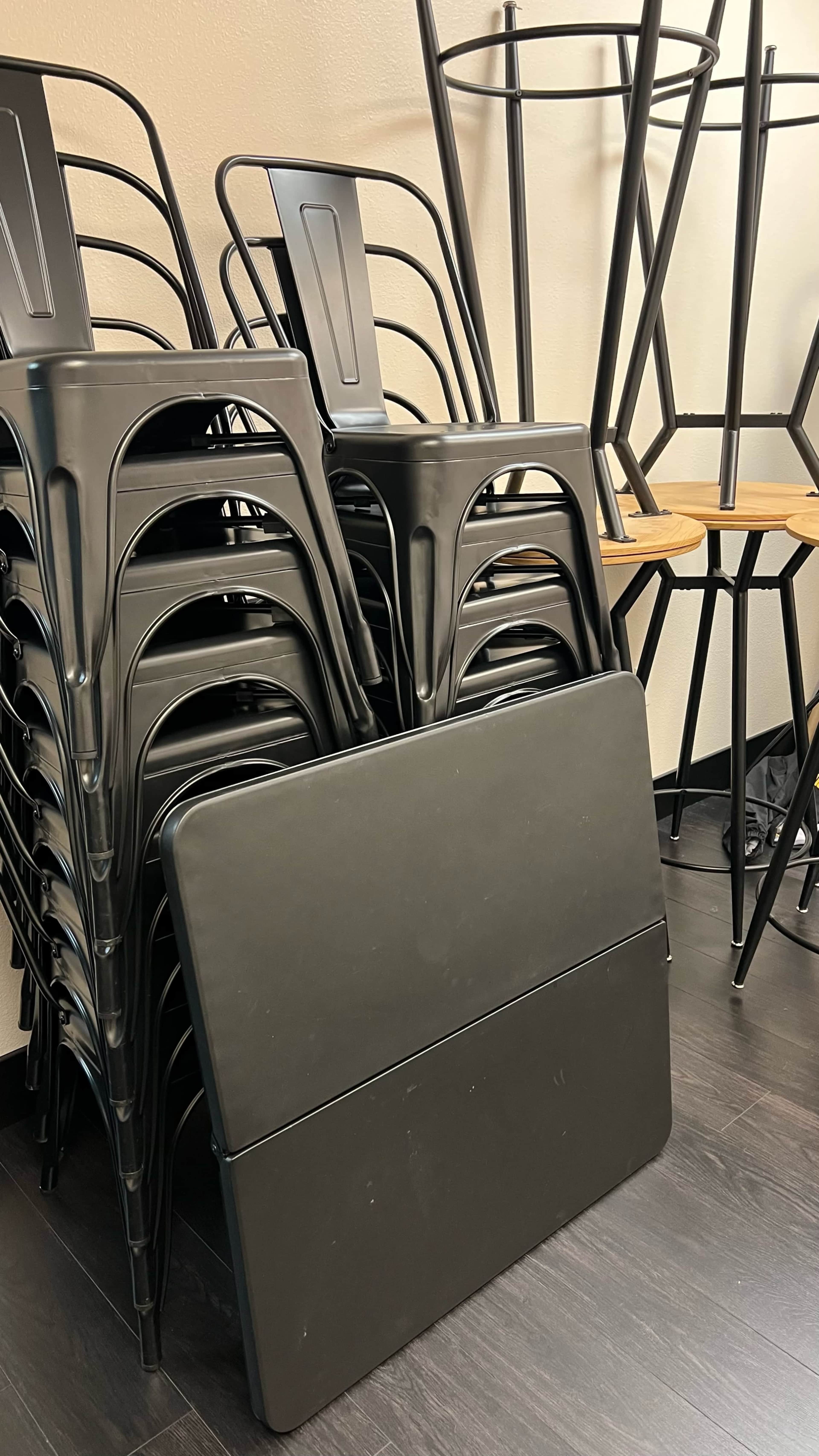 A stack of black metal chairs is leaning against a wall next to a folded black table in a room with wooden bar stools.
