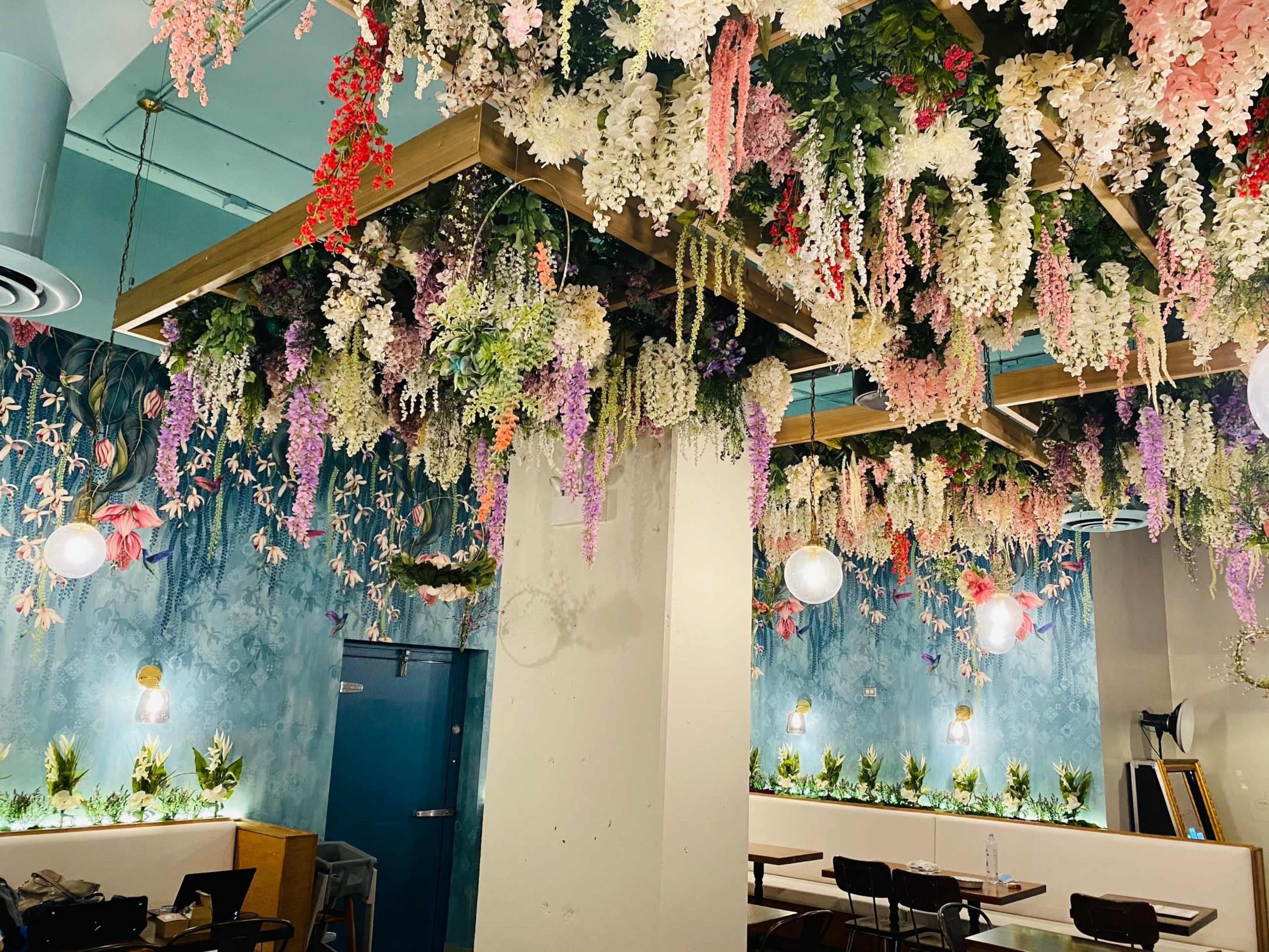 The image shows a cafe interior adorned with hanging artificial flowers and greenery, along with light fixtures and a blue wall behind wooden seating areas.