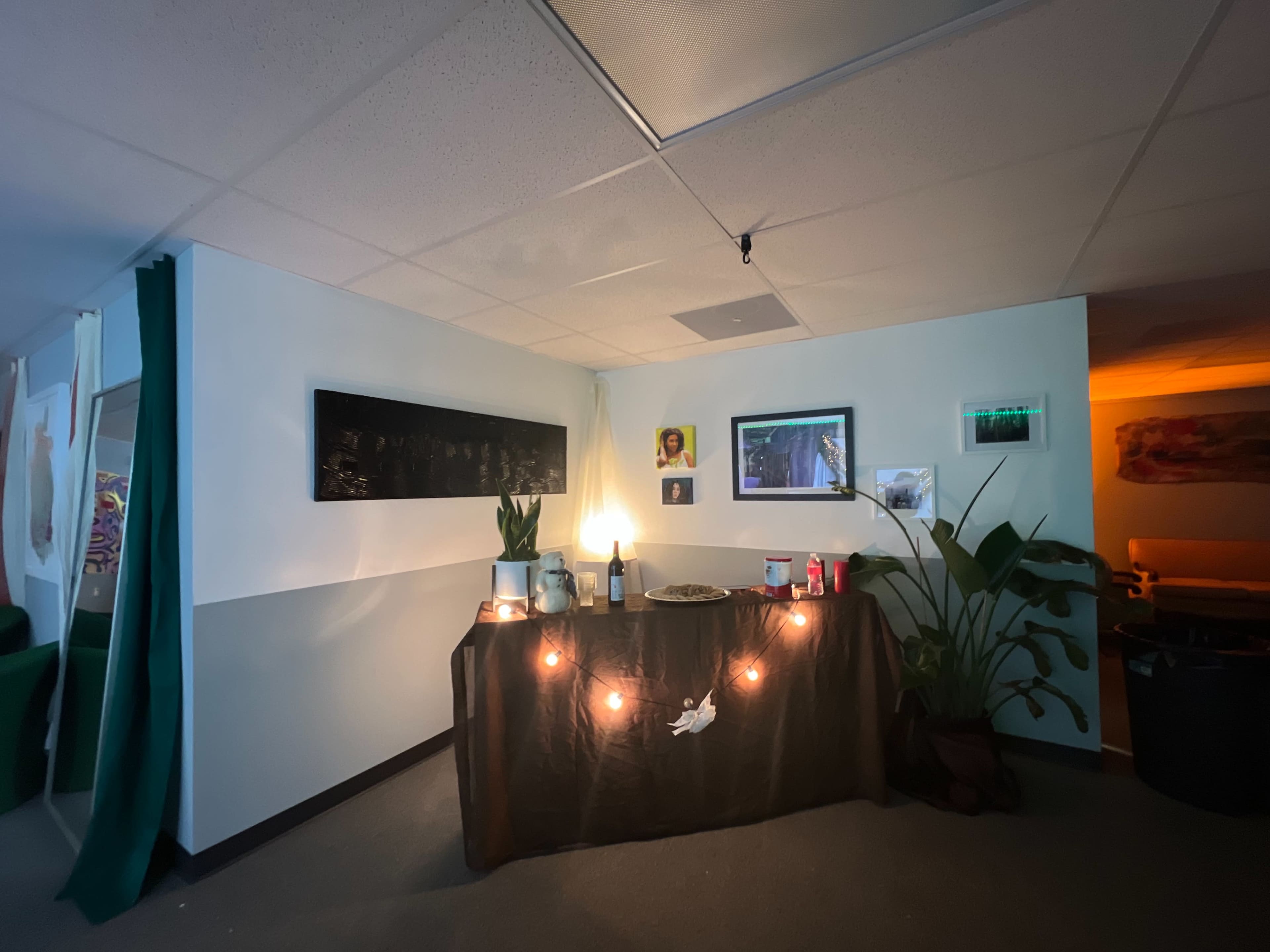 A corner of a room features a table draped in dark fabric, adorned with a lamp, plants, and various photographs, with string lights positioned around the display.