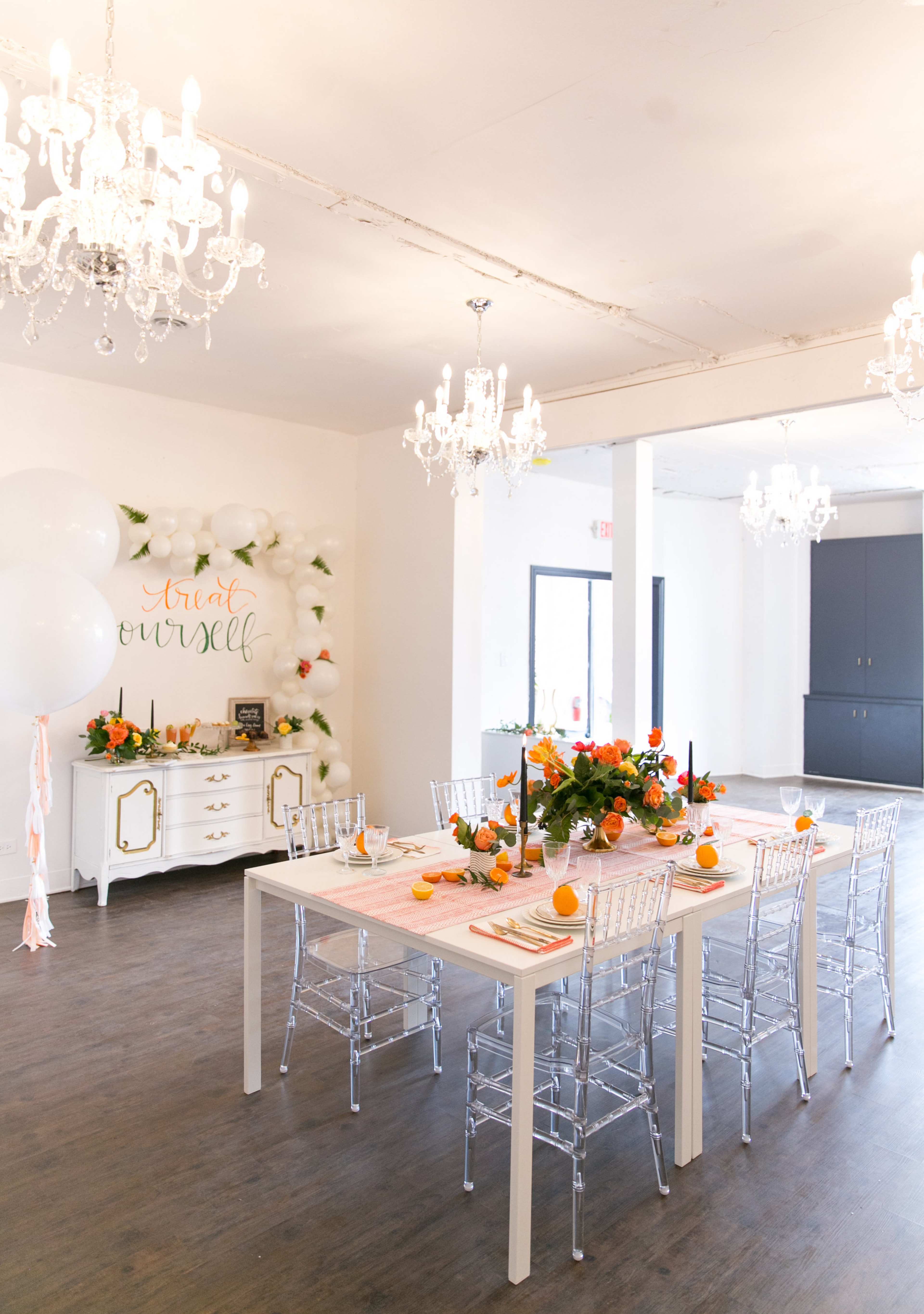 A long dining table is set with decorative flowers and fruits, surrounded by clear plastic chairs, in a room with chandeliers and an elegant wall display.