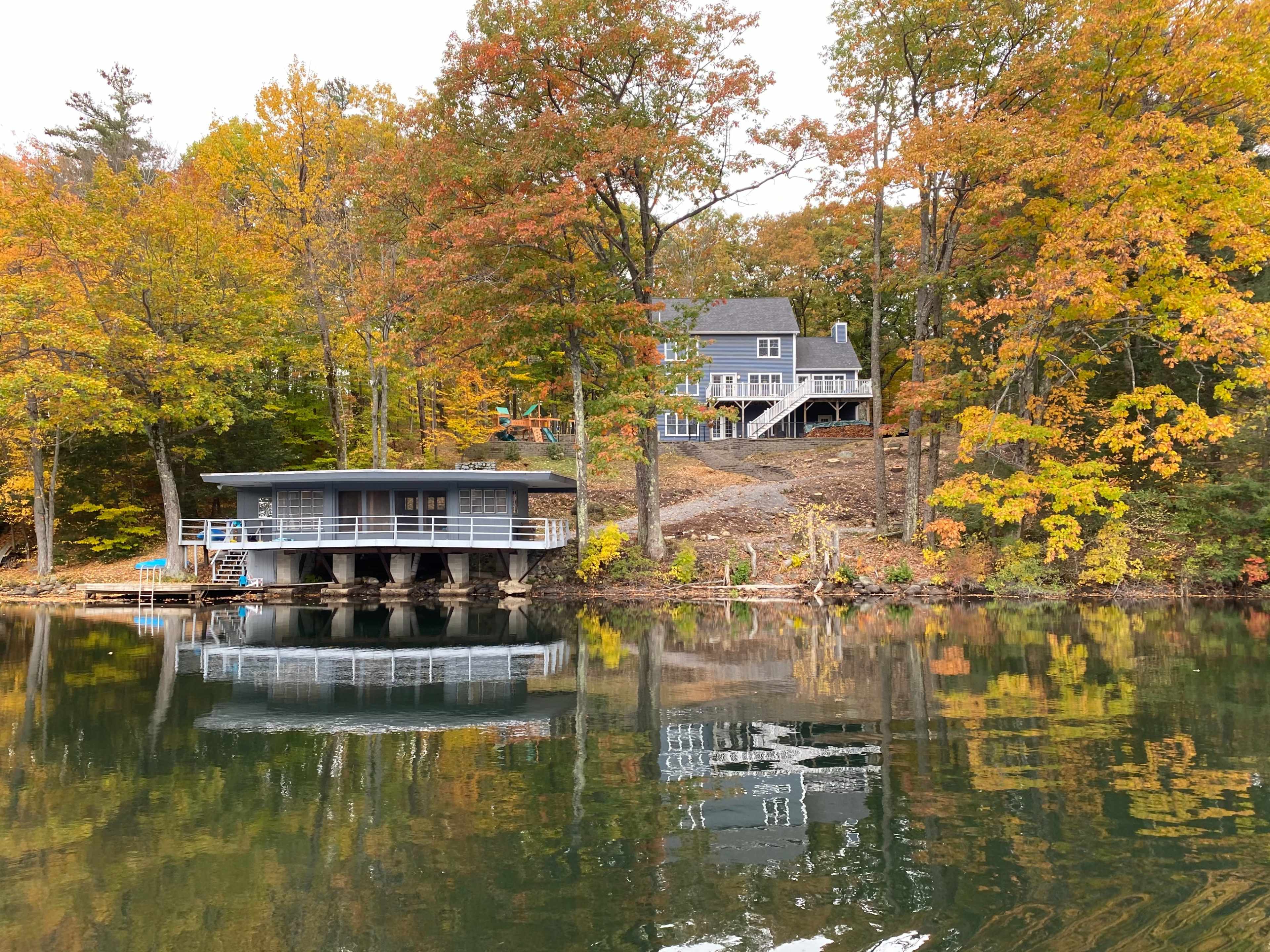 A serene lakeside scene features two houses with autumn foliage reflecting on the water's surface.