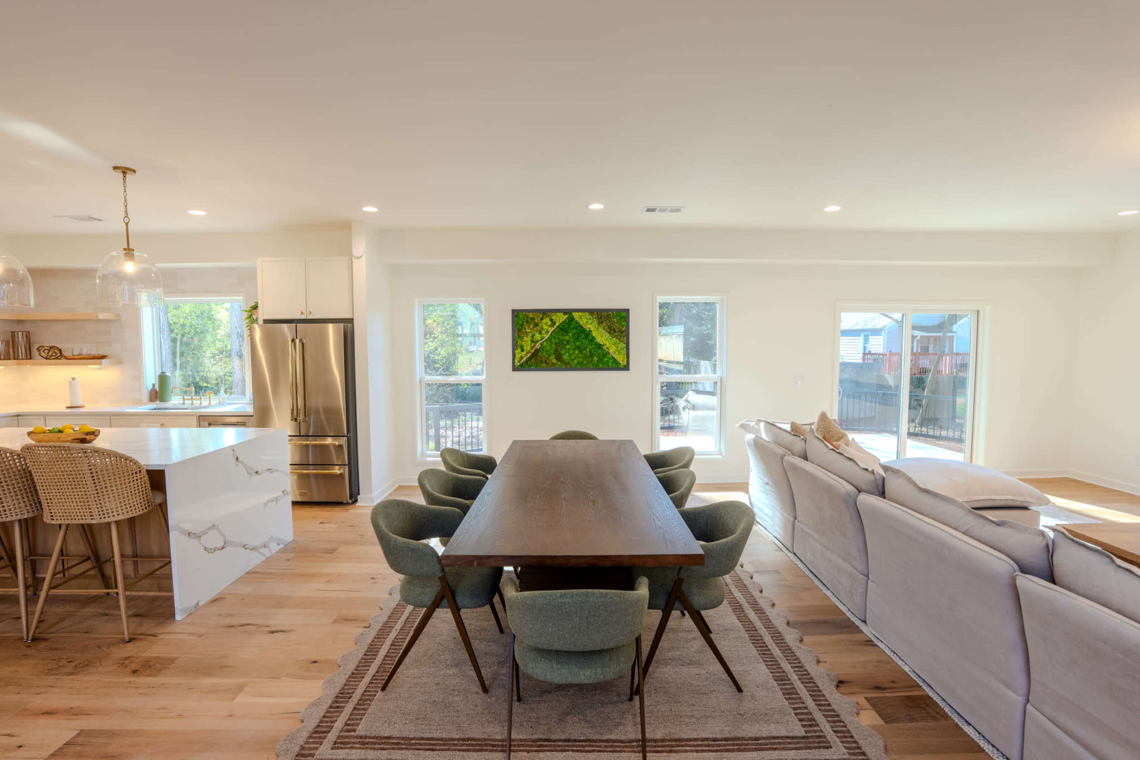 The image shows a modern open-concept living space featuring a dining table with green chairs, a kitchen with stainless steel appliances, and a comfortable seating area with a light-colored sofa.