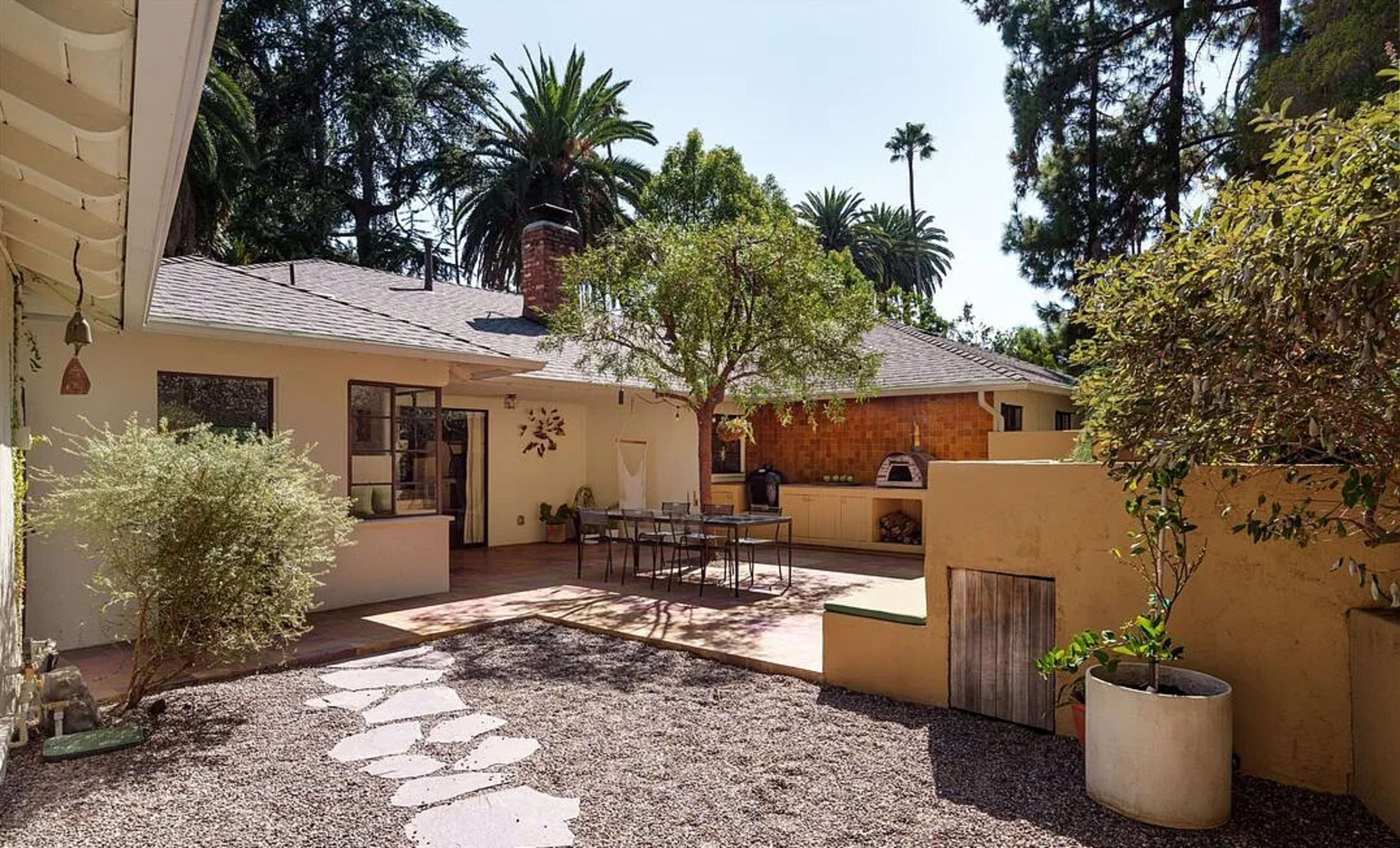 The image shows a spacious outdoor area of a home featuring a patio with a dining table, surrounded by well-maintained greenery and palm trees.