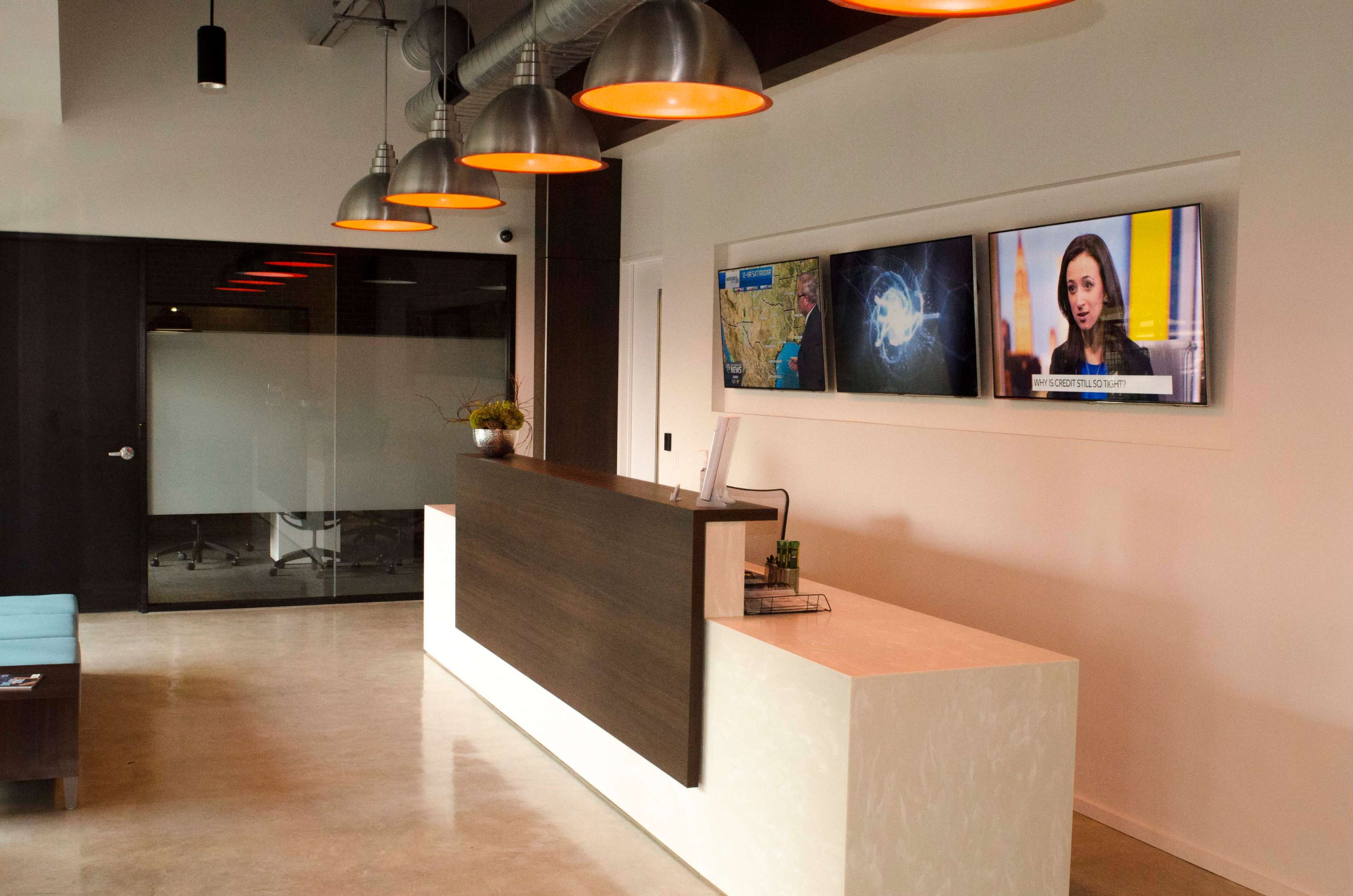 A modern reception area features a white front desk, two large screens displaying information and news, and overhead pendant lighting.