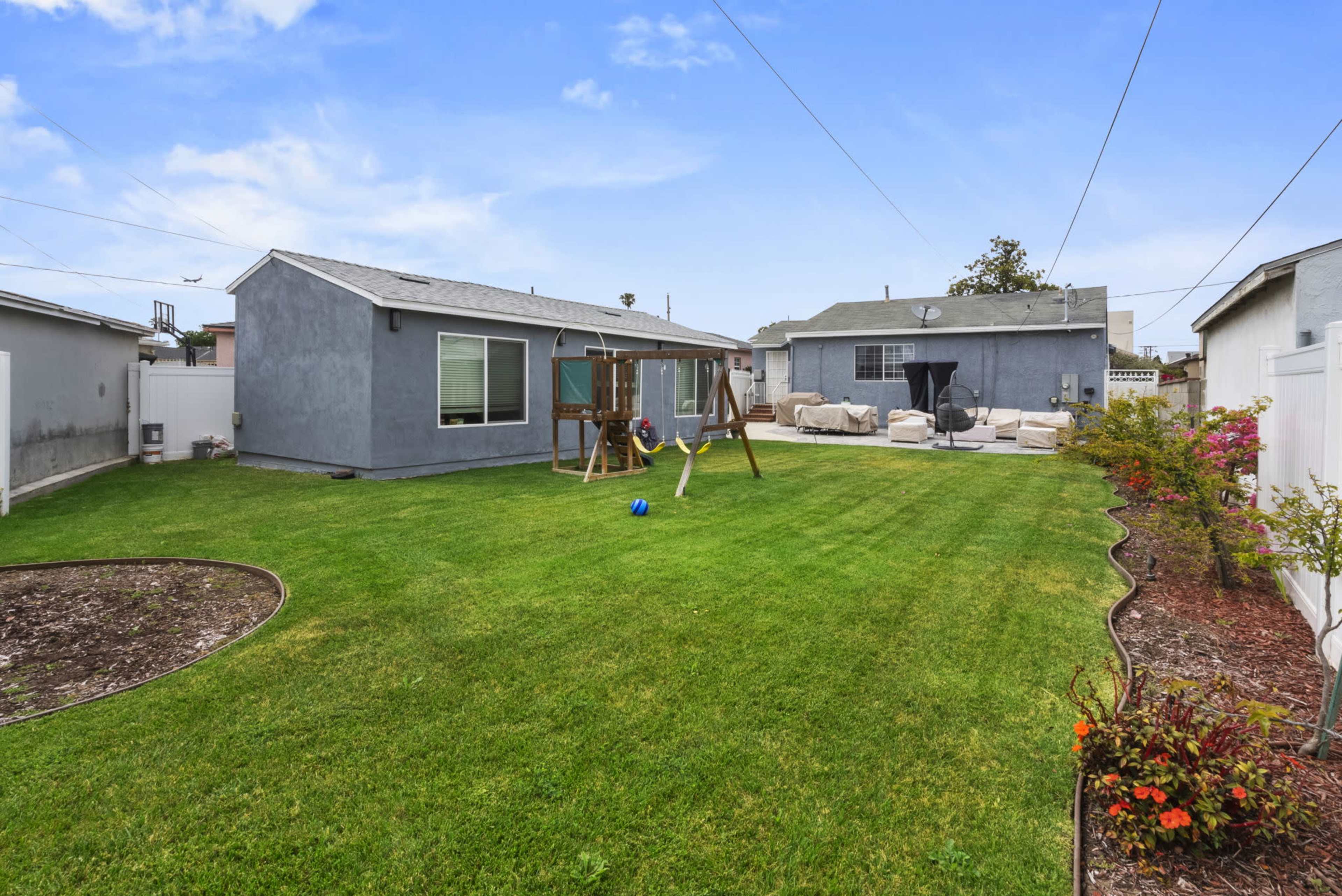 The image shows a spacious backyard with well-maintained grass, a playground set, patio furniture, and surrounding houses.