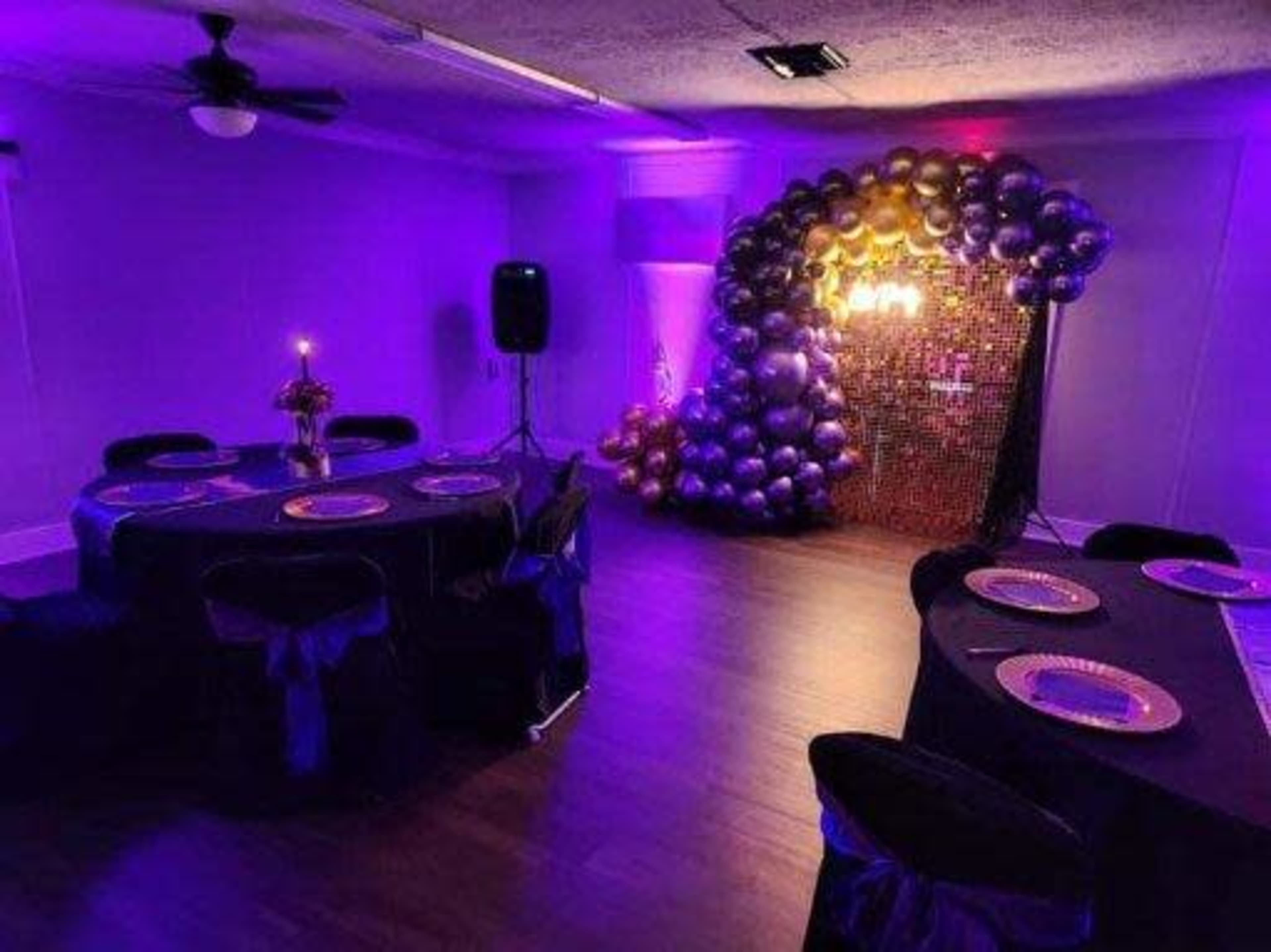 A decorated room features wooden flooring with two tables set with plates and a large arch of balloons in purple and gold against a backdrop of fairy lights.