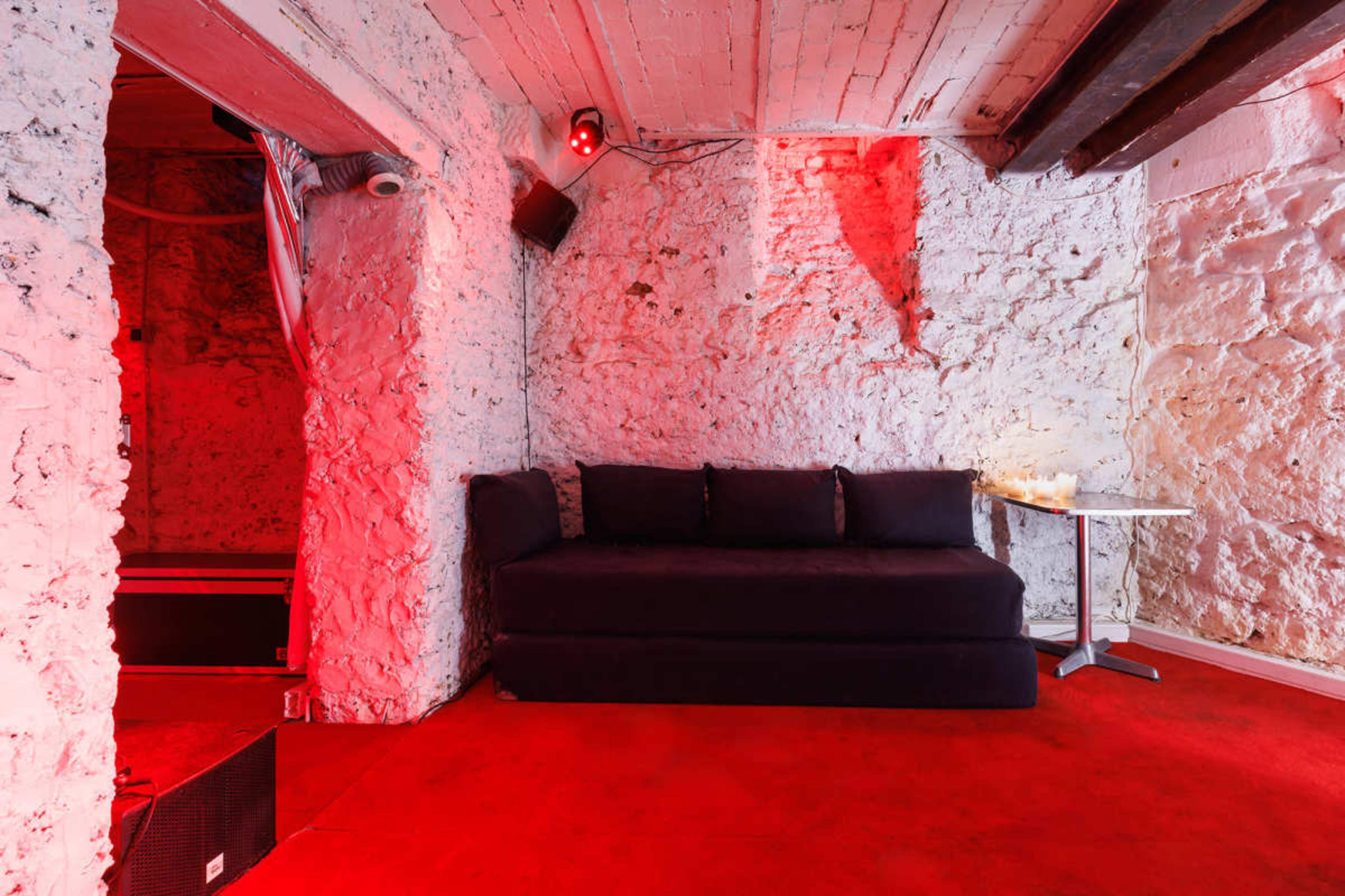 A small, dimly lit room with stone walls features a dark couch and a side table on red carpeting.