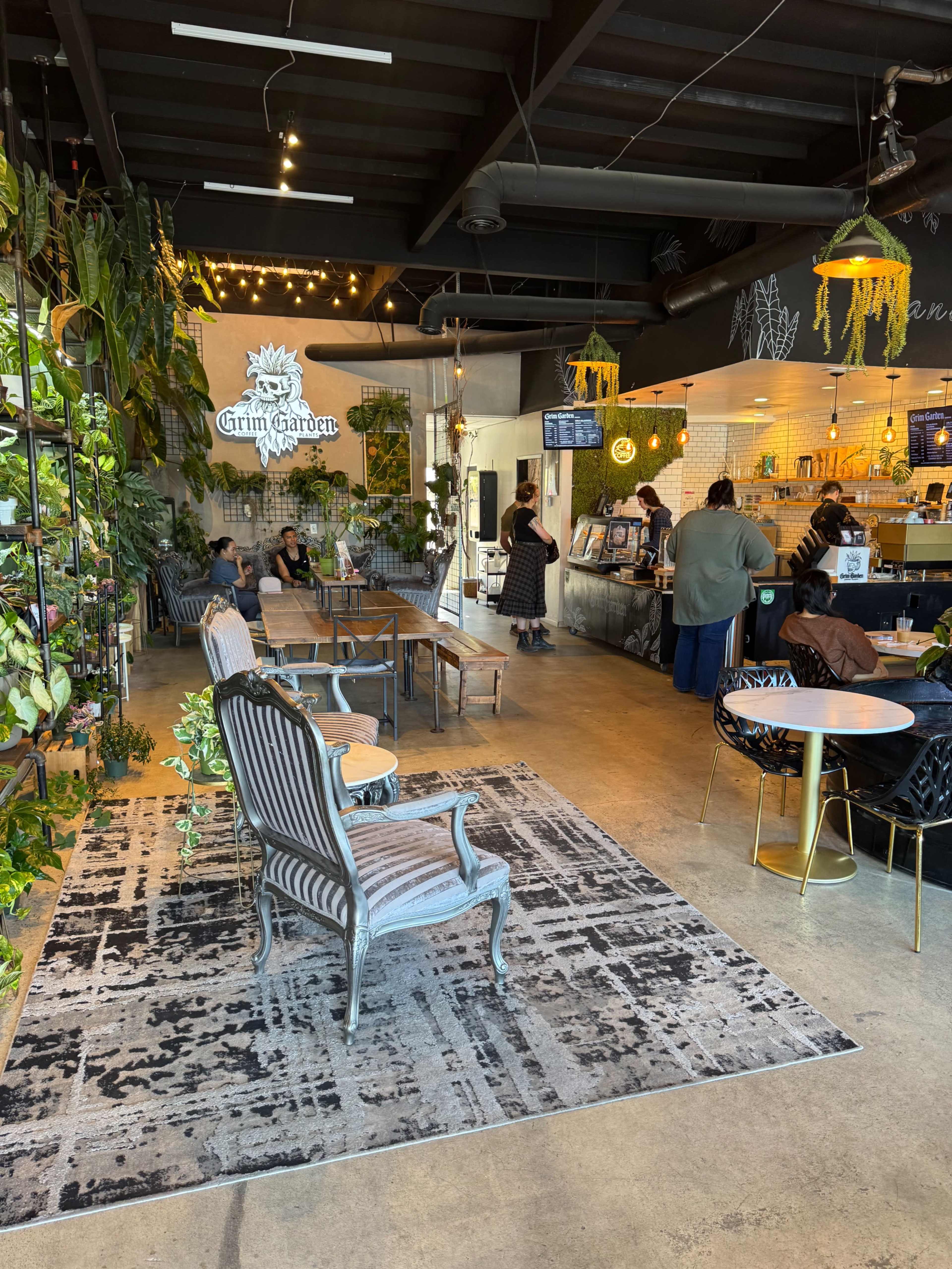 The image shows a cozy café interior featuring greenery, a counter with baristas, and several patrons seated at tables.