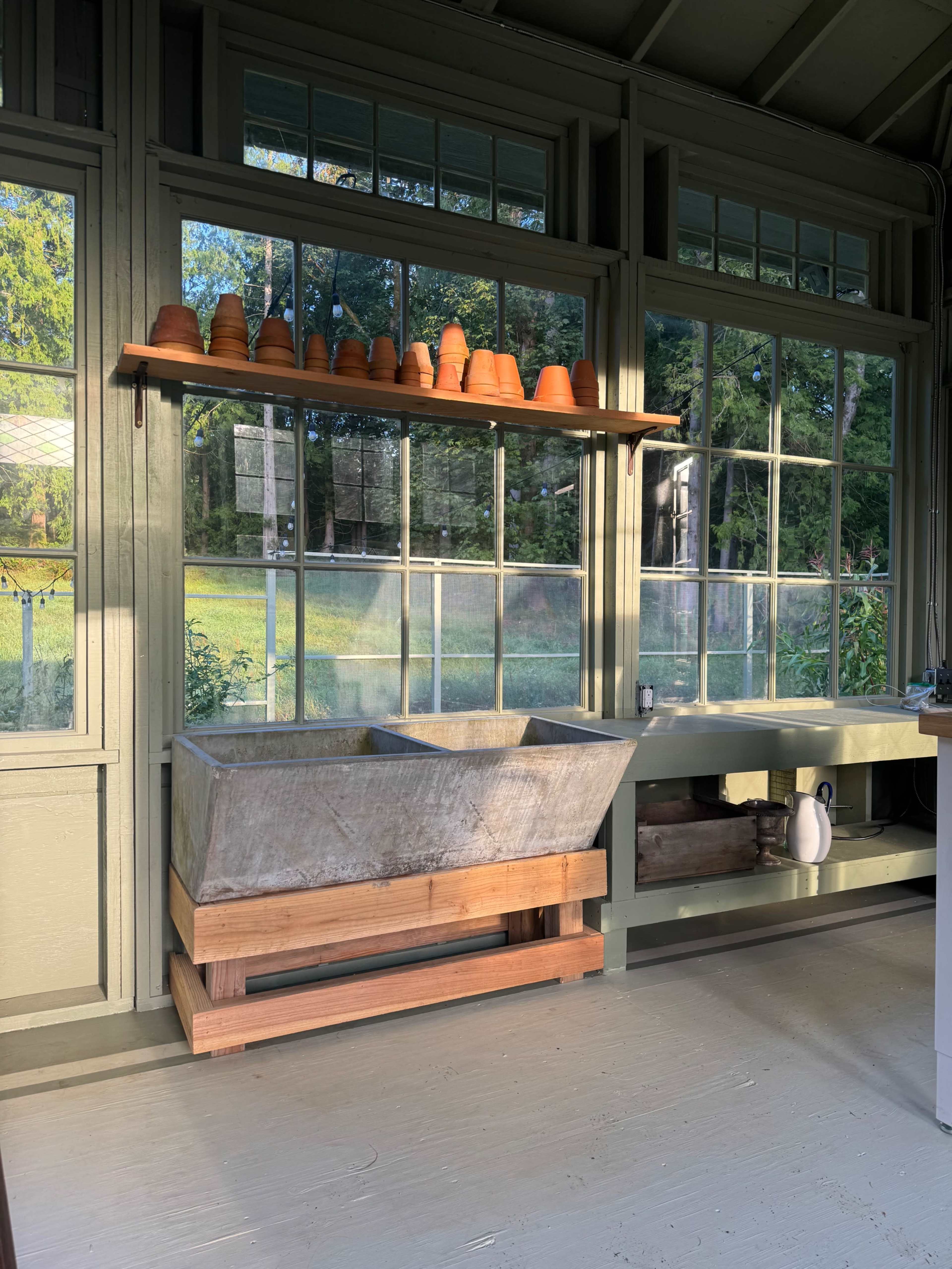 The image shows a rustic wash basin made of concrete perched on a wooden frame, positioned beneath a shelf lined with clay pots and surrounded by large windows letting in natural light.