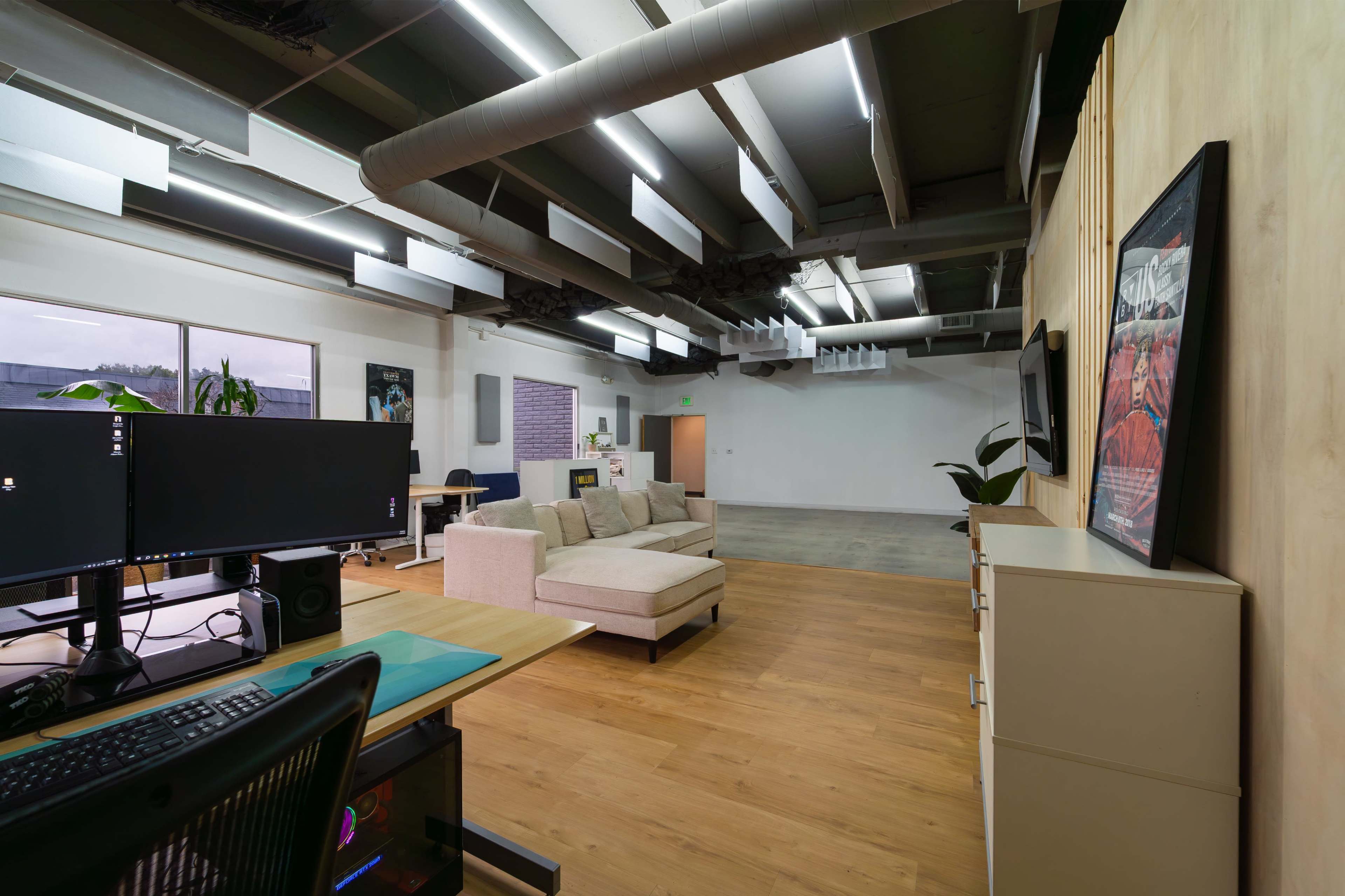 The image shows a spacious room with a lounge area featuring a light-colored sectional sofa, a desk with computer monitors, and wooden flooring.