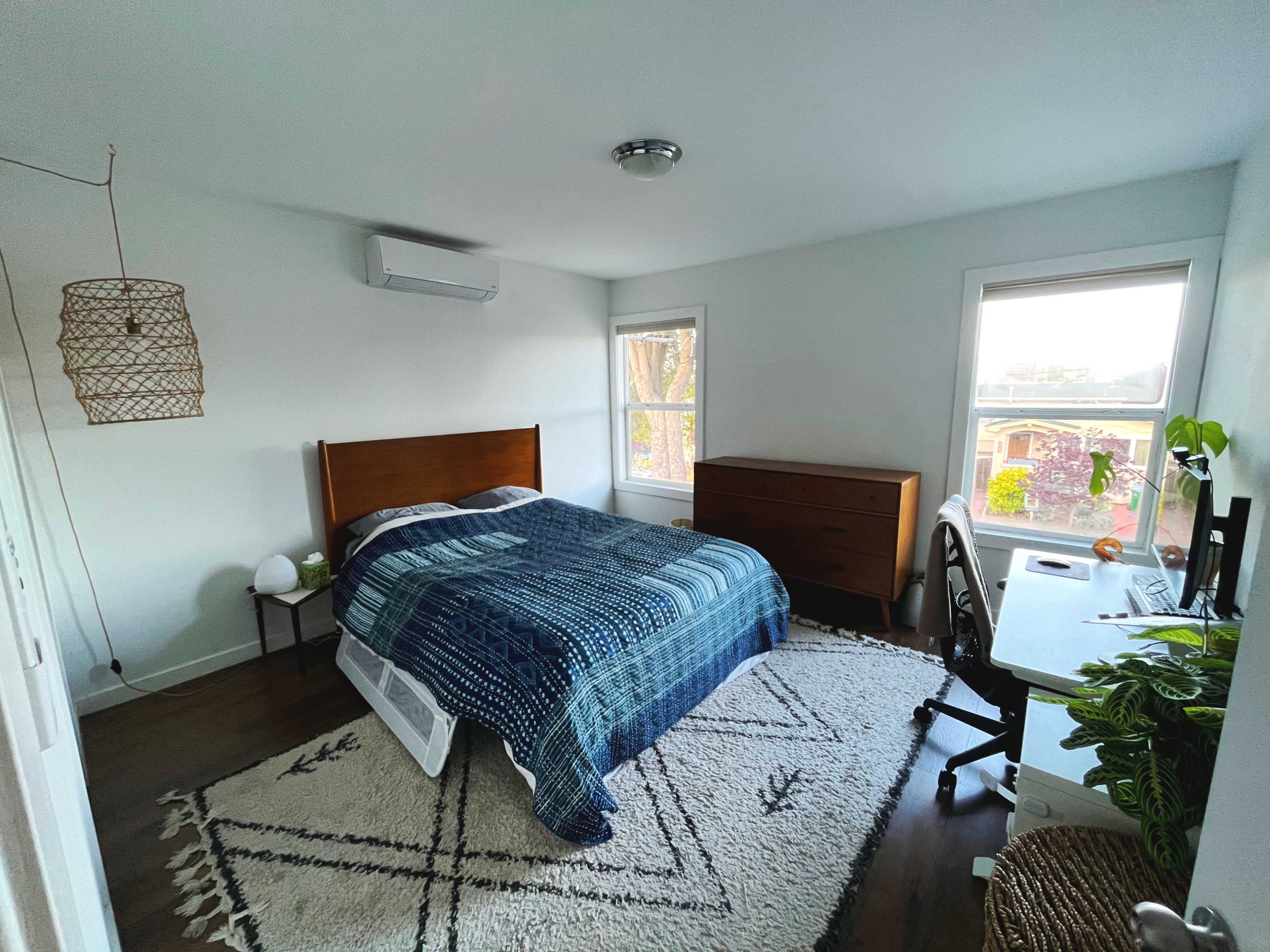 The image shows a bedroom featuring a wooden bed, a dresser, a small bedside table, and a desk with a plant, all set against white walls and a rug.