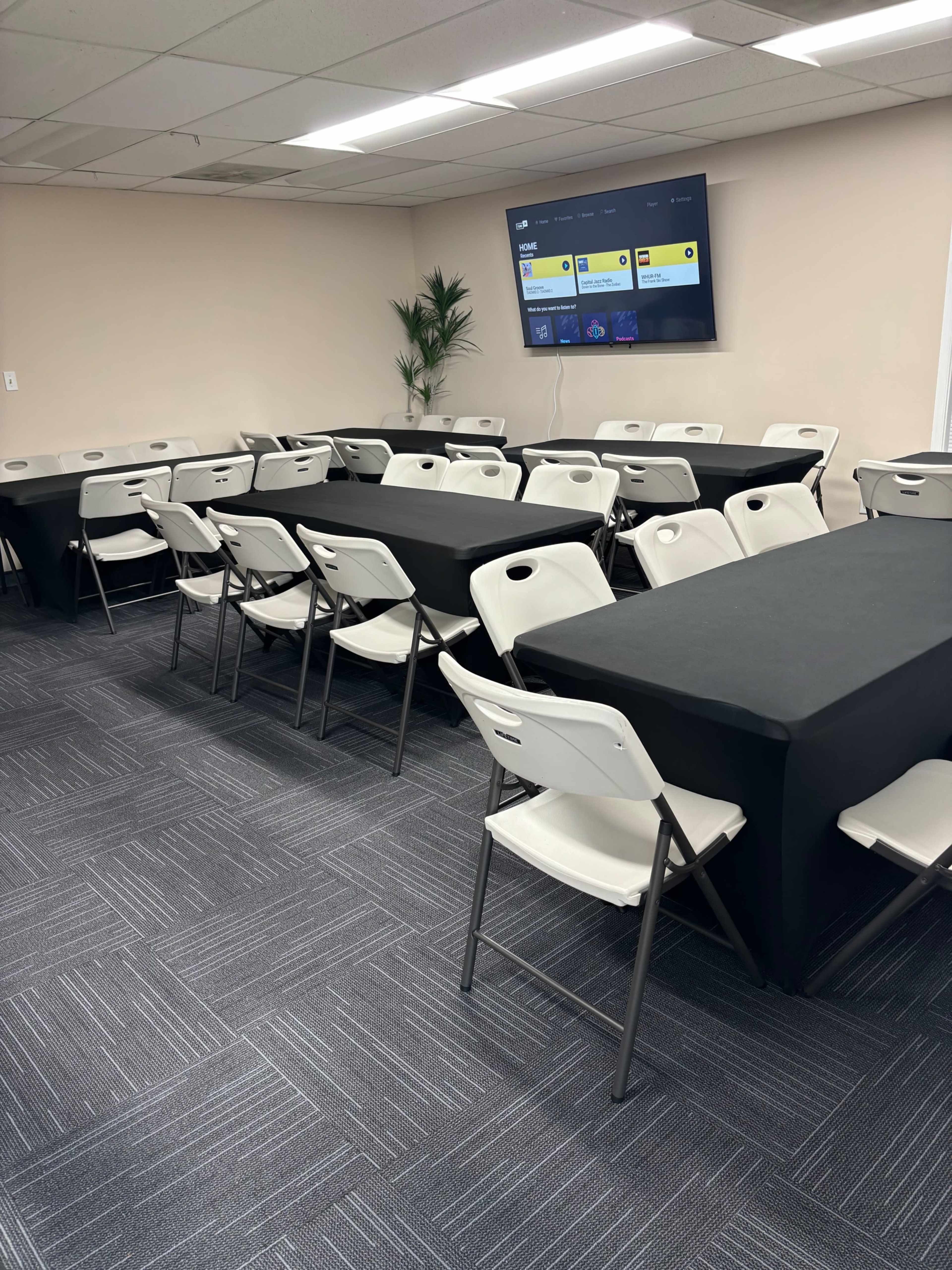 A conference room is set up with several rectangular tables covered in black tablecloths and surrounded by folding chairs, with a television mounted on the wall displaying a screen.