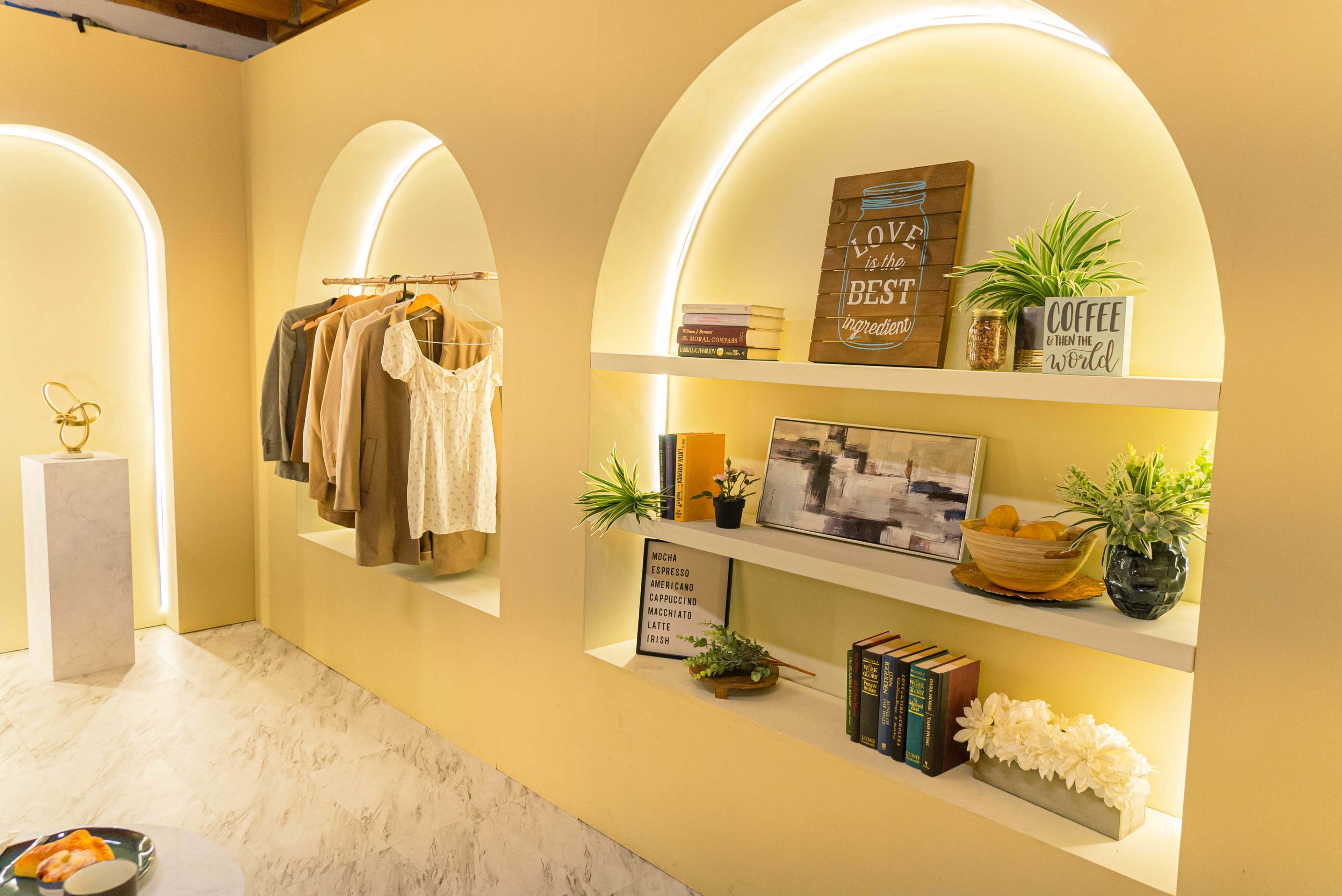 The scene features a stylish interior with illuminated archways, displaying neatly arranged clothing on hangers and decorative shelves filled with plants, books, and framed art.