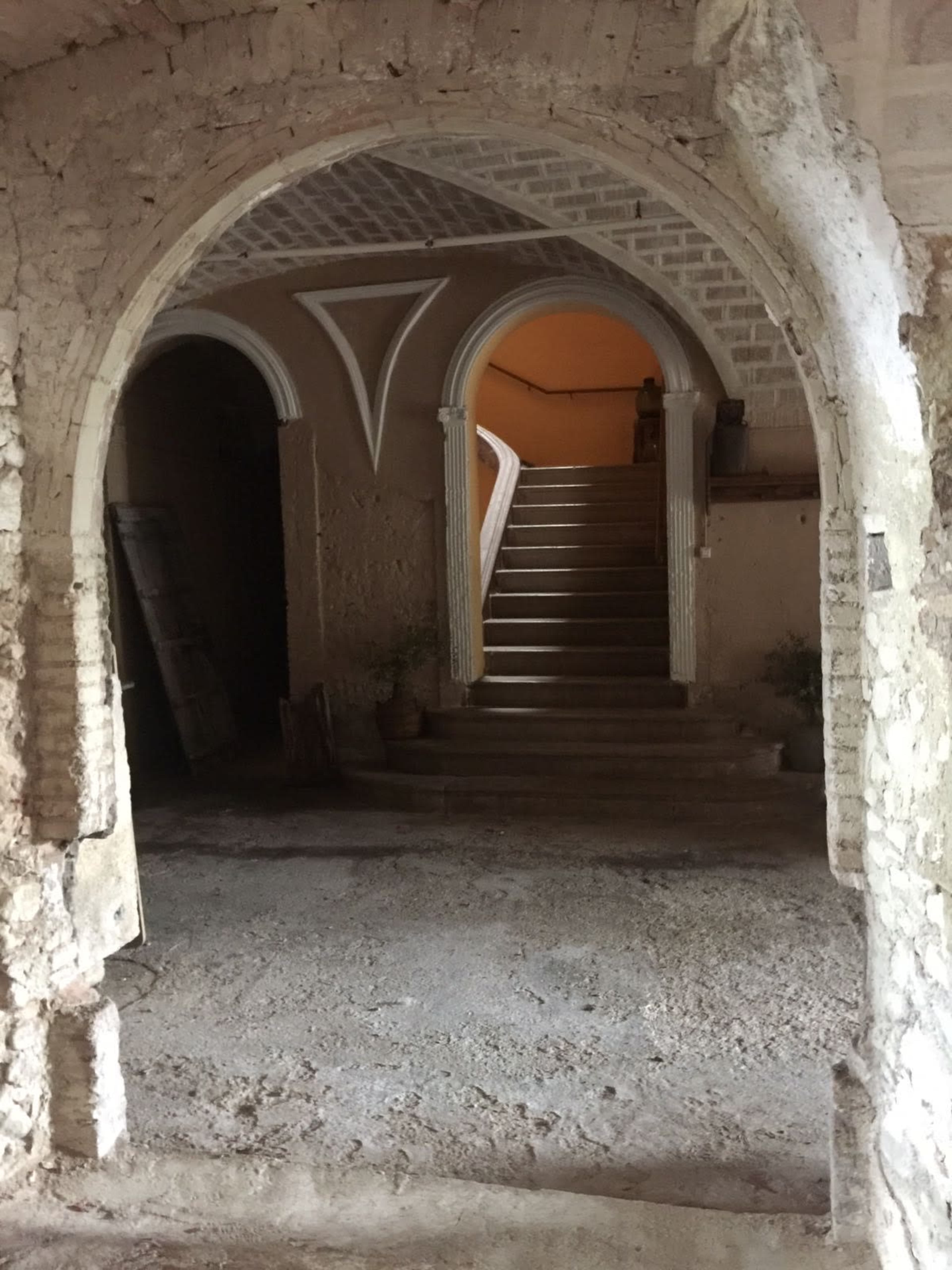 An old, unfinished interior space with stone arches leading to a staircase and walls displaying peeling paint.