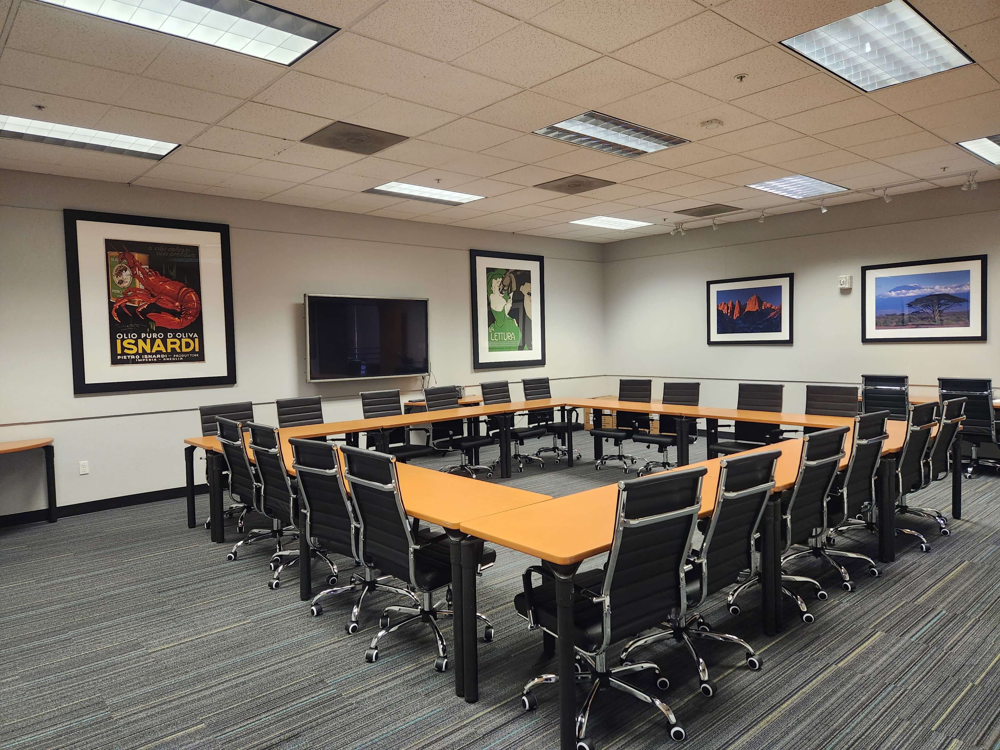 The image shows a spacious conference room arranged with a large, rectangular table surrounded by black ergonomic chairs, accompanied by framed artwork on the walls.