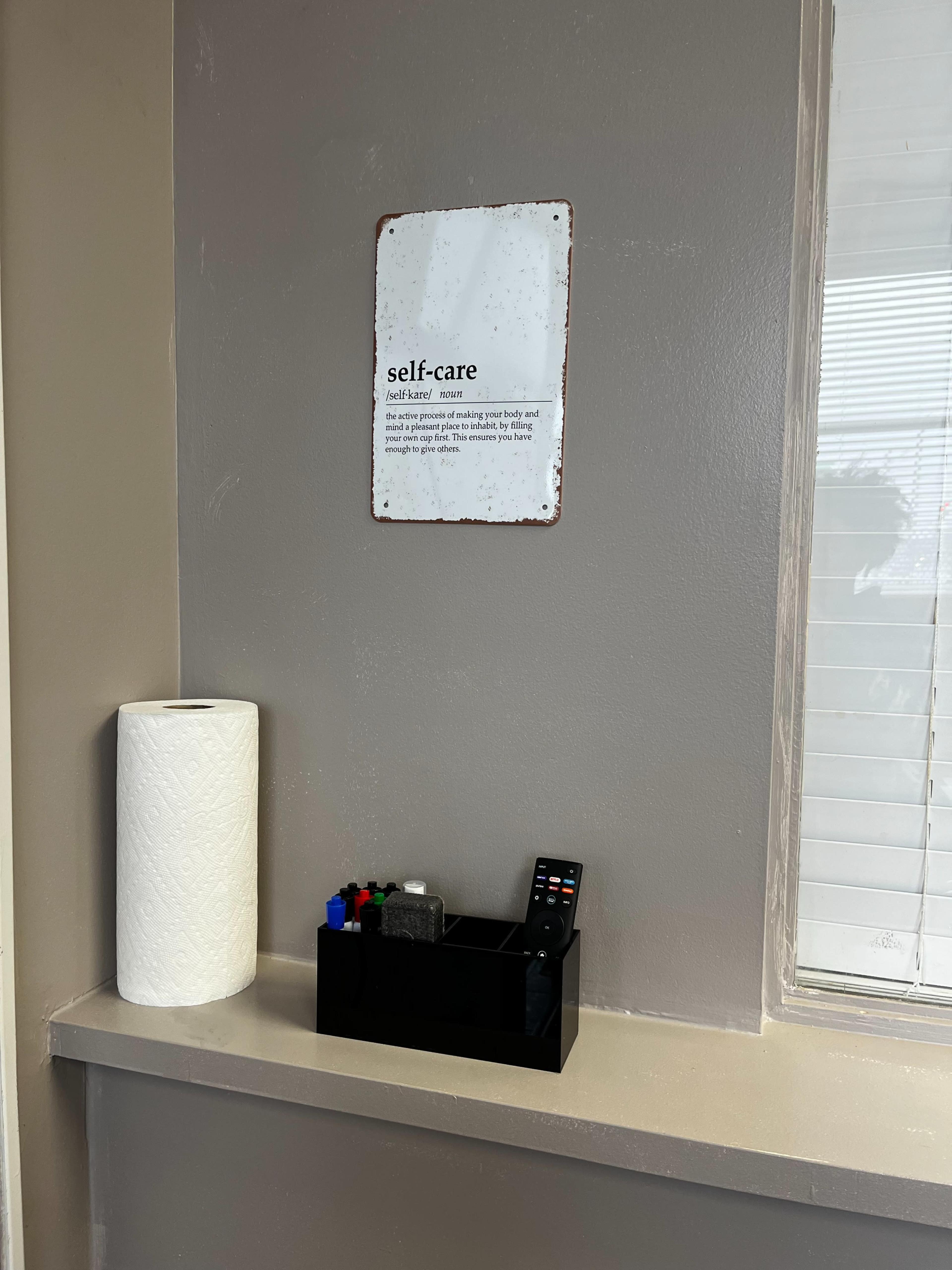 A wall with a self-care sign mounted above a shelf that holds a black organizer with a remote control and miscellaneous items, alongside a roll of paper towels.