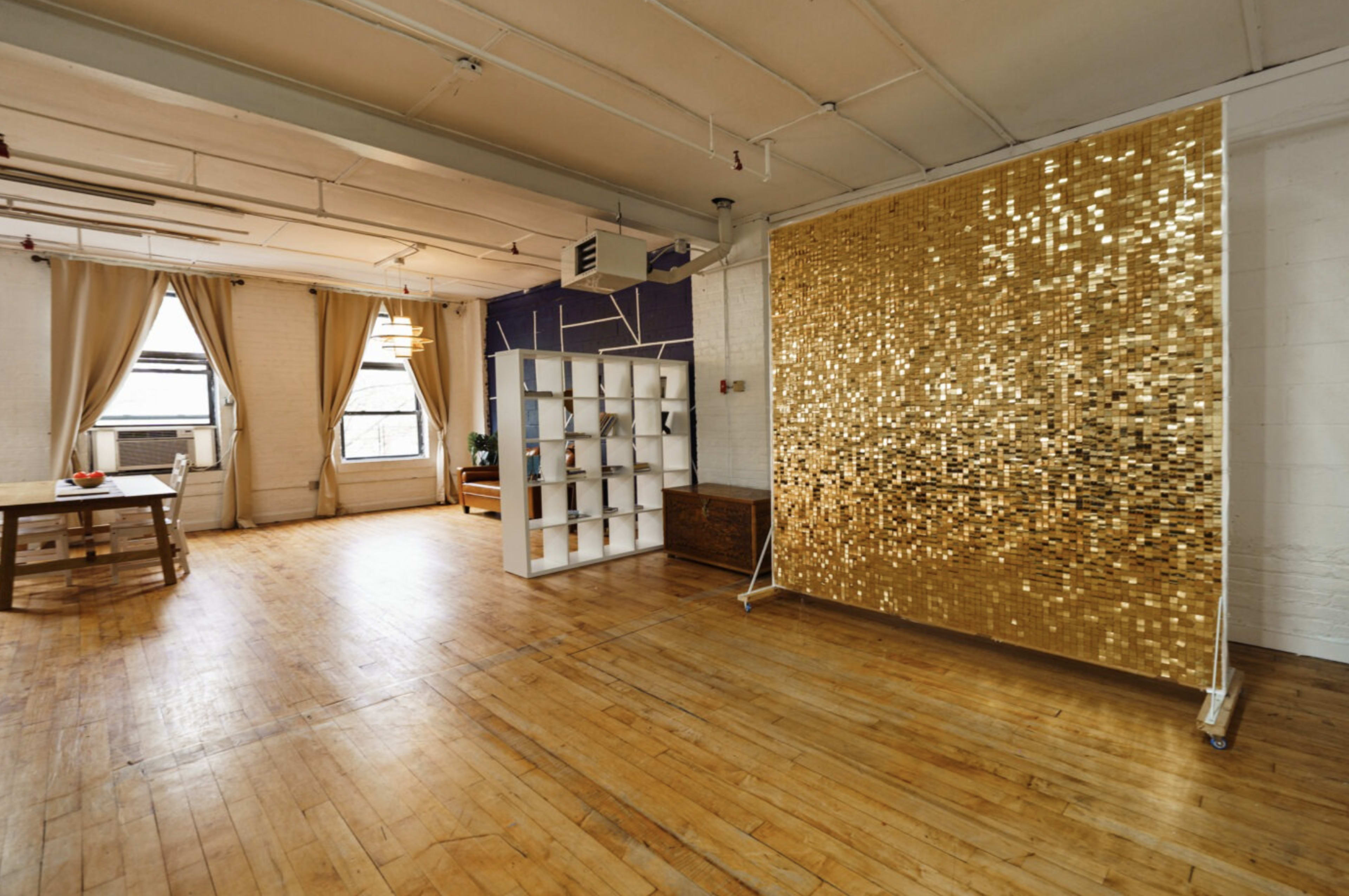 A spacious room with wooden floors, a large gold decorative wall, a white shelving unit, and a wooden table set for dining.