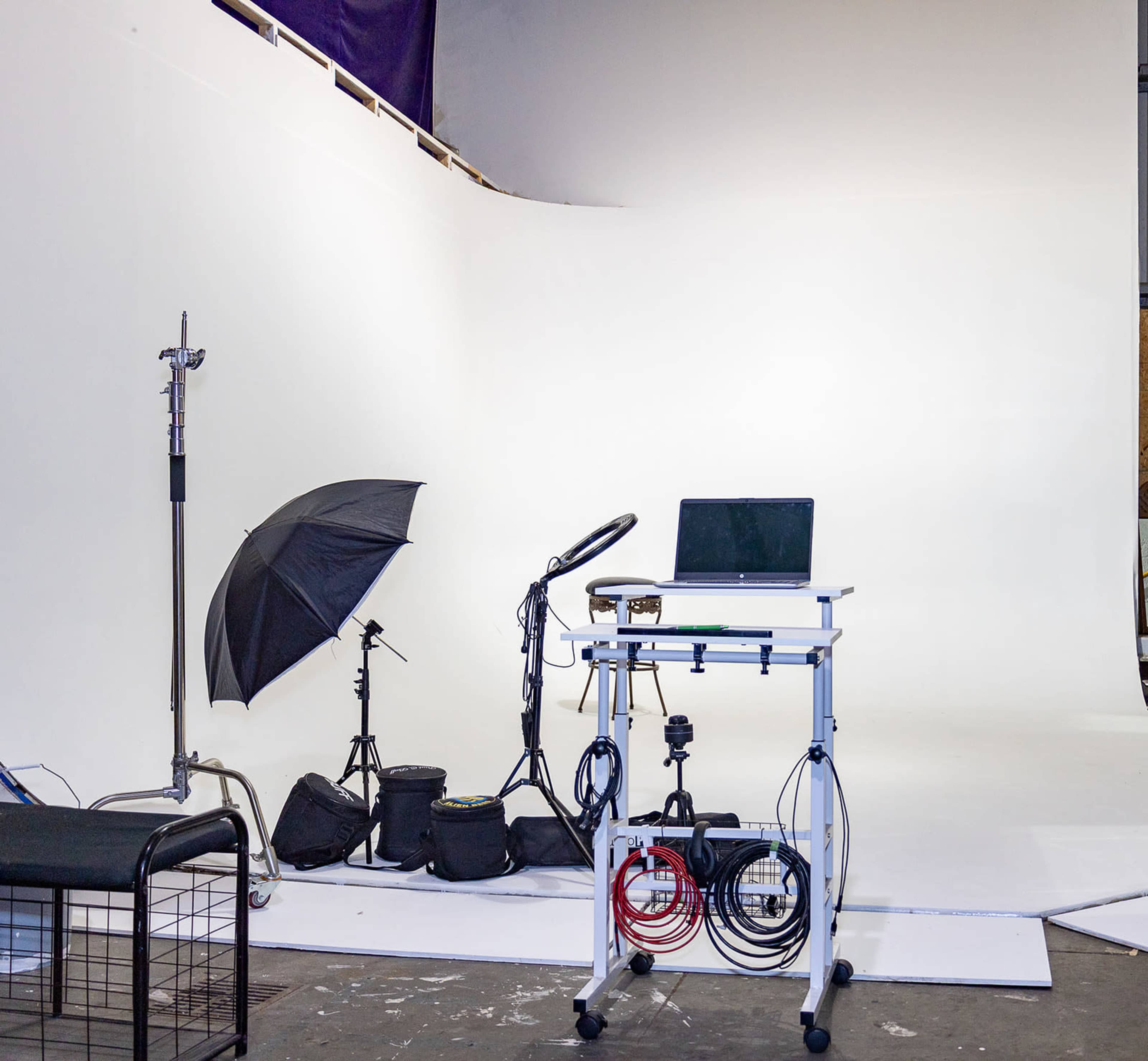 The image showcases a photography studio with a blank white backdrop, a table with a laptop, and various lighting equipment set up.