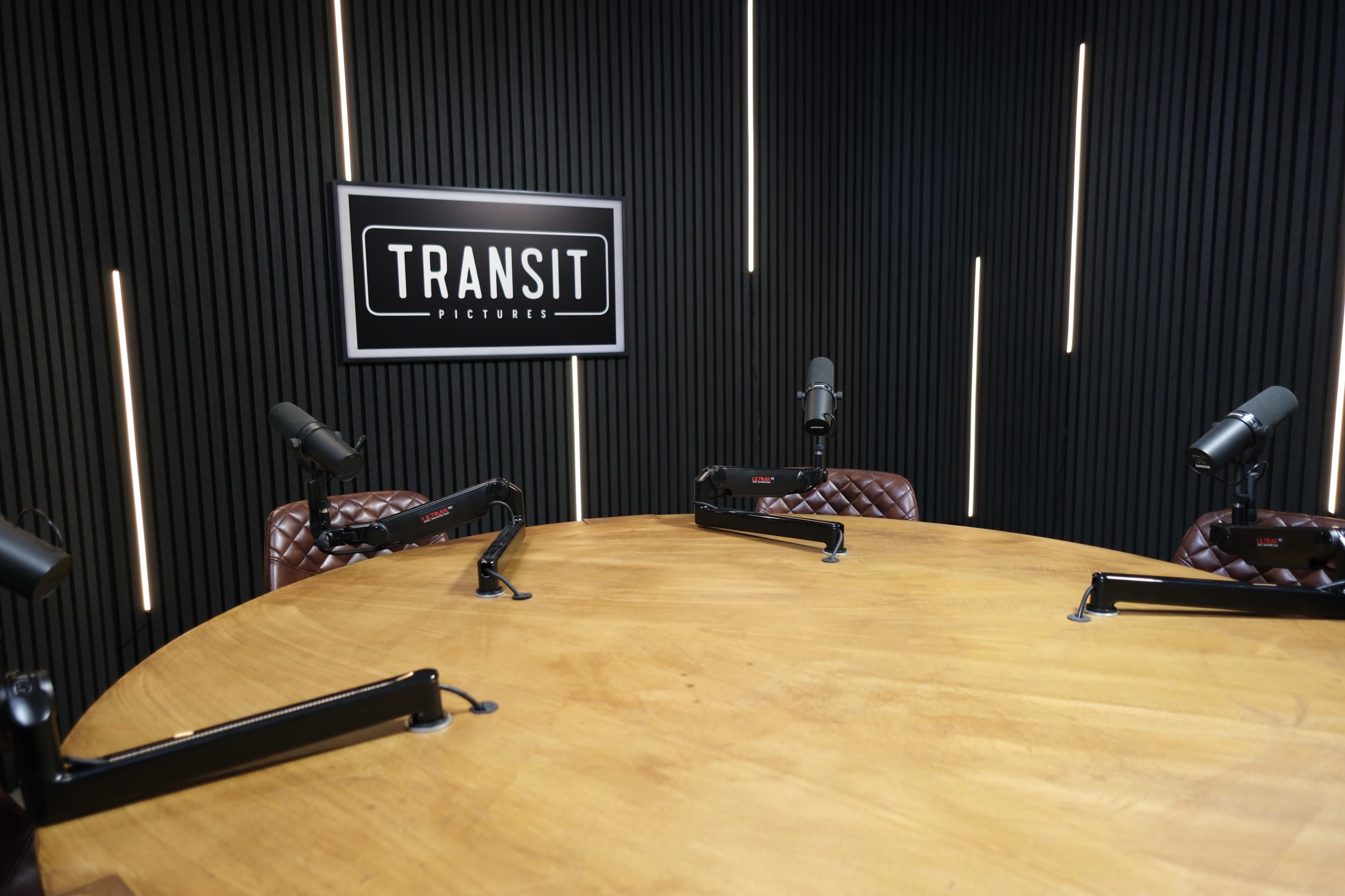 The image shows a podcast studio featuring a large wooden table surrounded by three microphone stands, with a wall display that reads "TRANSIT PICTURES."