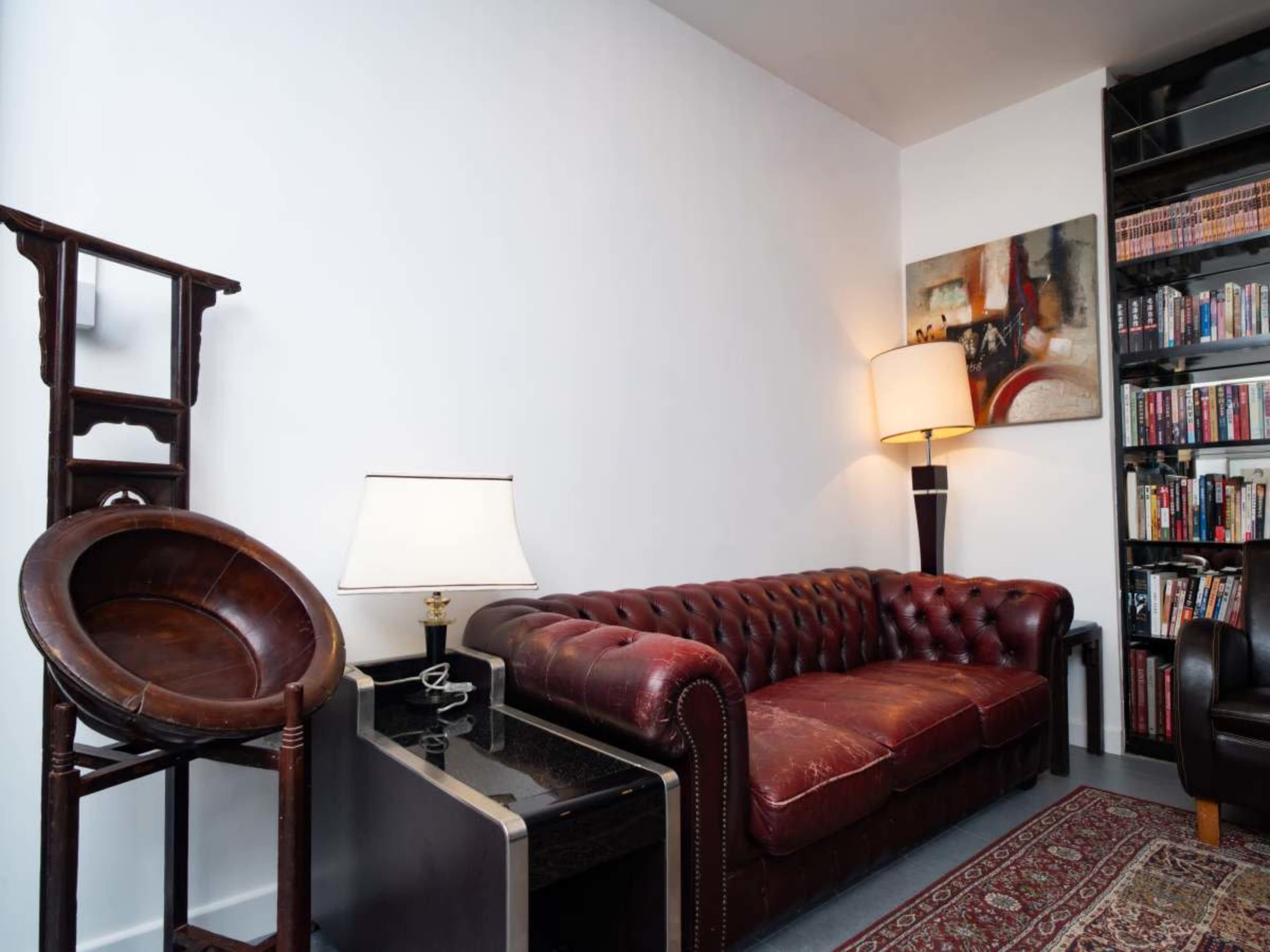 The image shows a chesterfield sofa in a room with a lamp, a small table, a decorative chair, and a bookshelf filled with books.