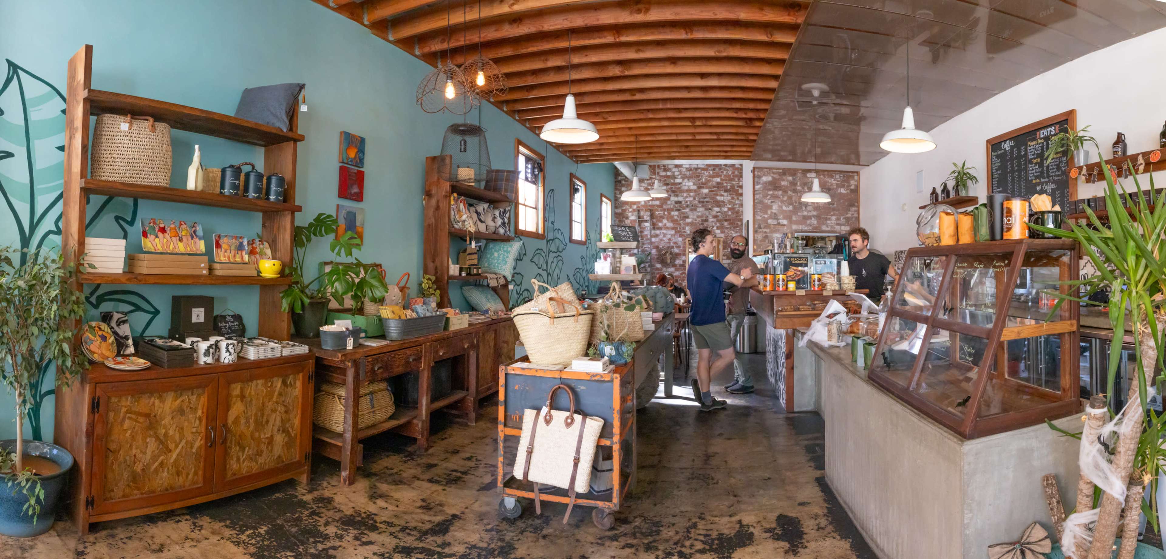 A cozy café interior features wooden shelves filled with various items, a counter where staff are serving customers, and a brick wall in the background.