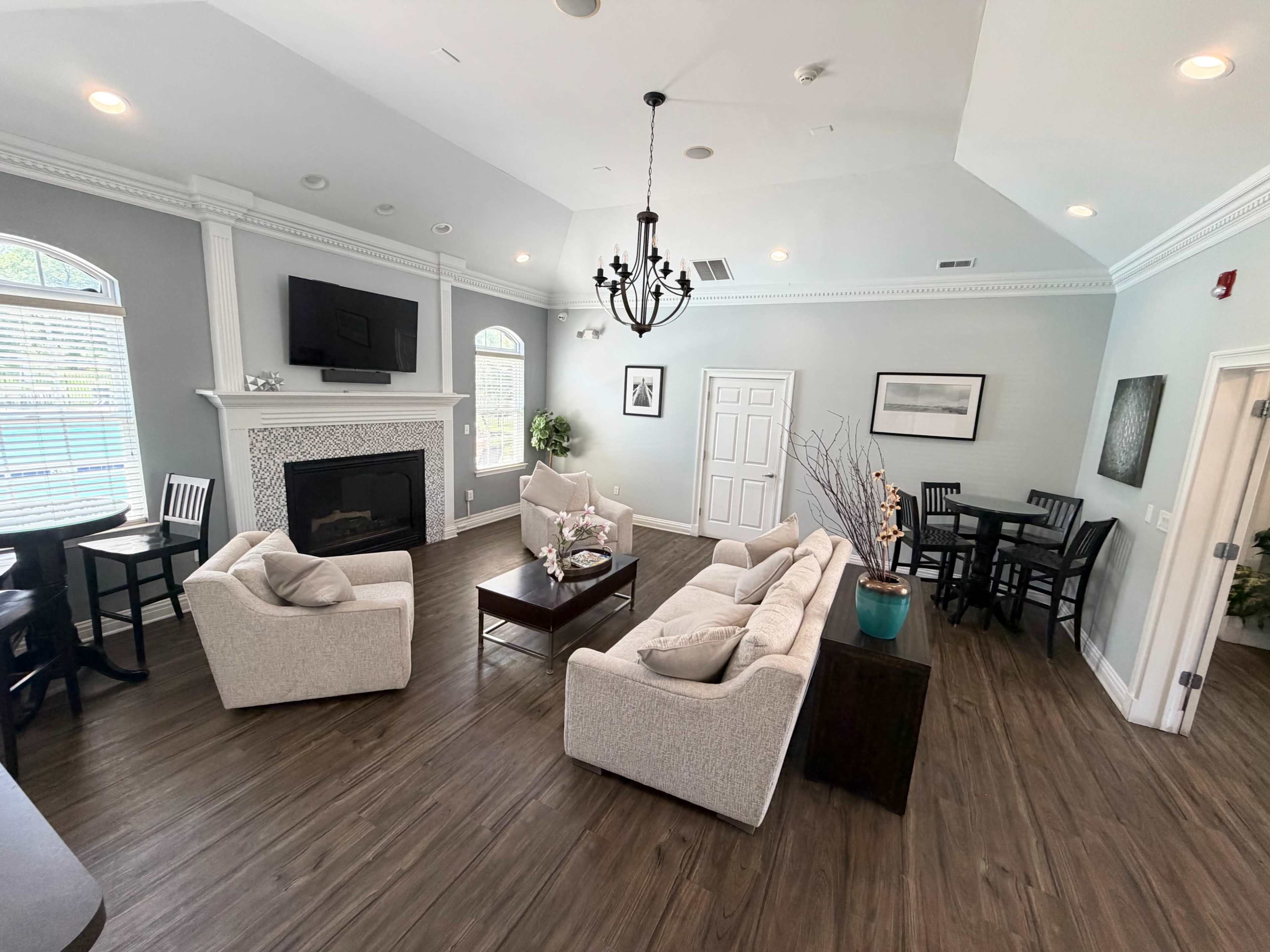 The image shows a modern lounge area with two beige sofas, a dark wooden coffee table, a fireplace, and a dining table set for four, all under a high ceiling with decorative molding.