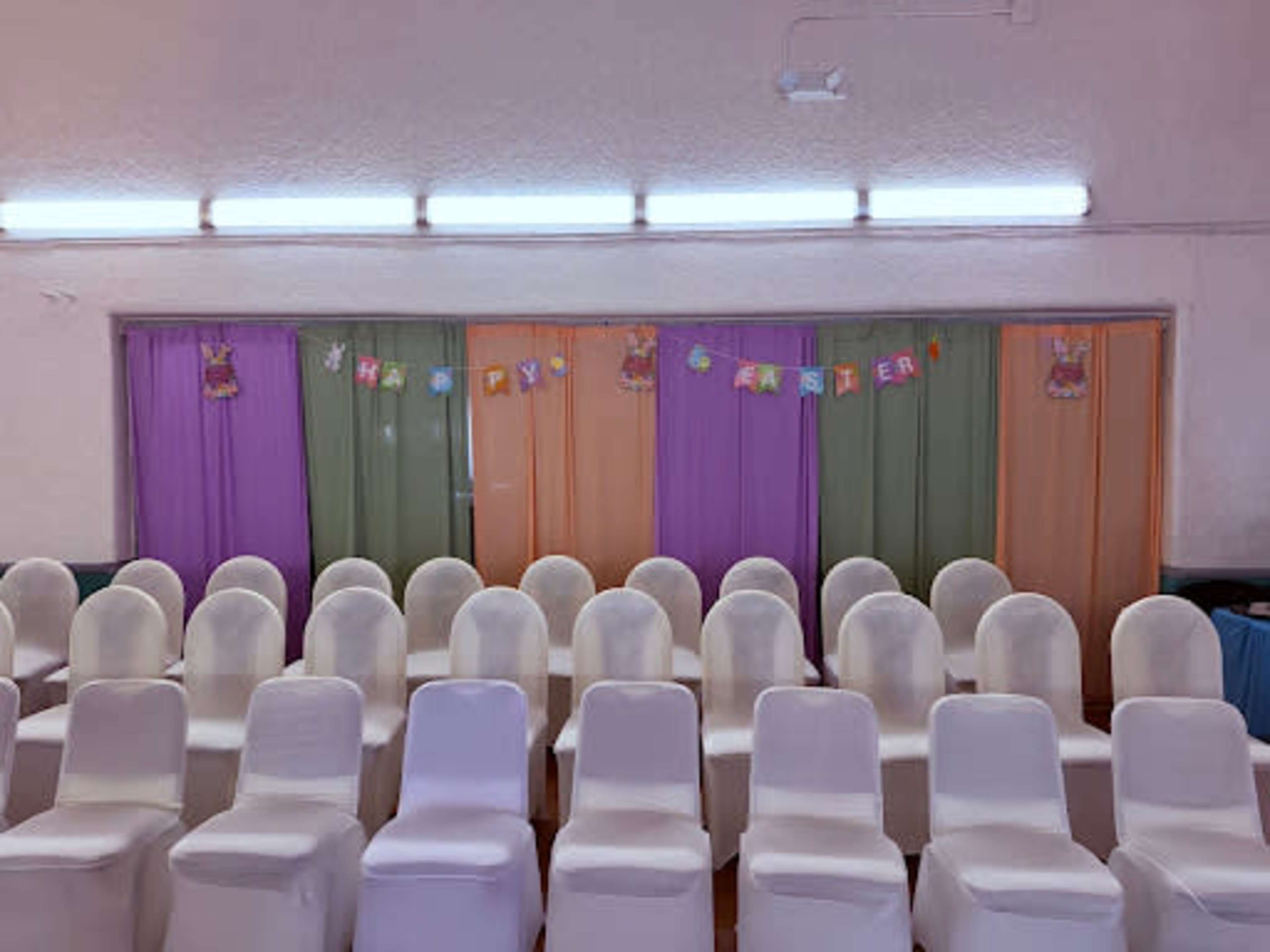 The image shows a row of white chairs arranged in a hall with colorful curtains and a decorative banner that reads "Happy Starter."