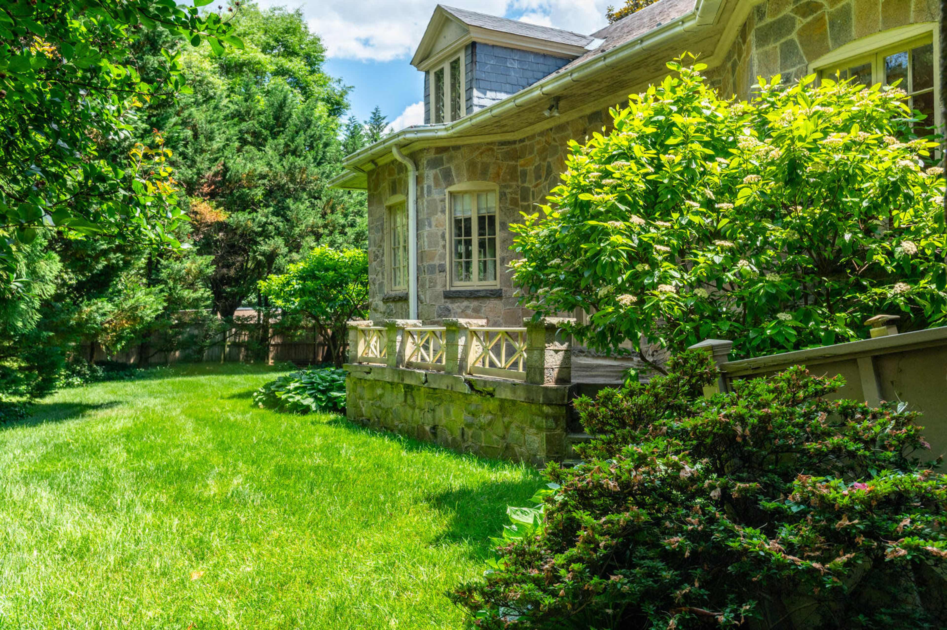 A stone house is surrounded by lush greenery and well-maintained grass in a shaded yard.