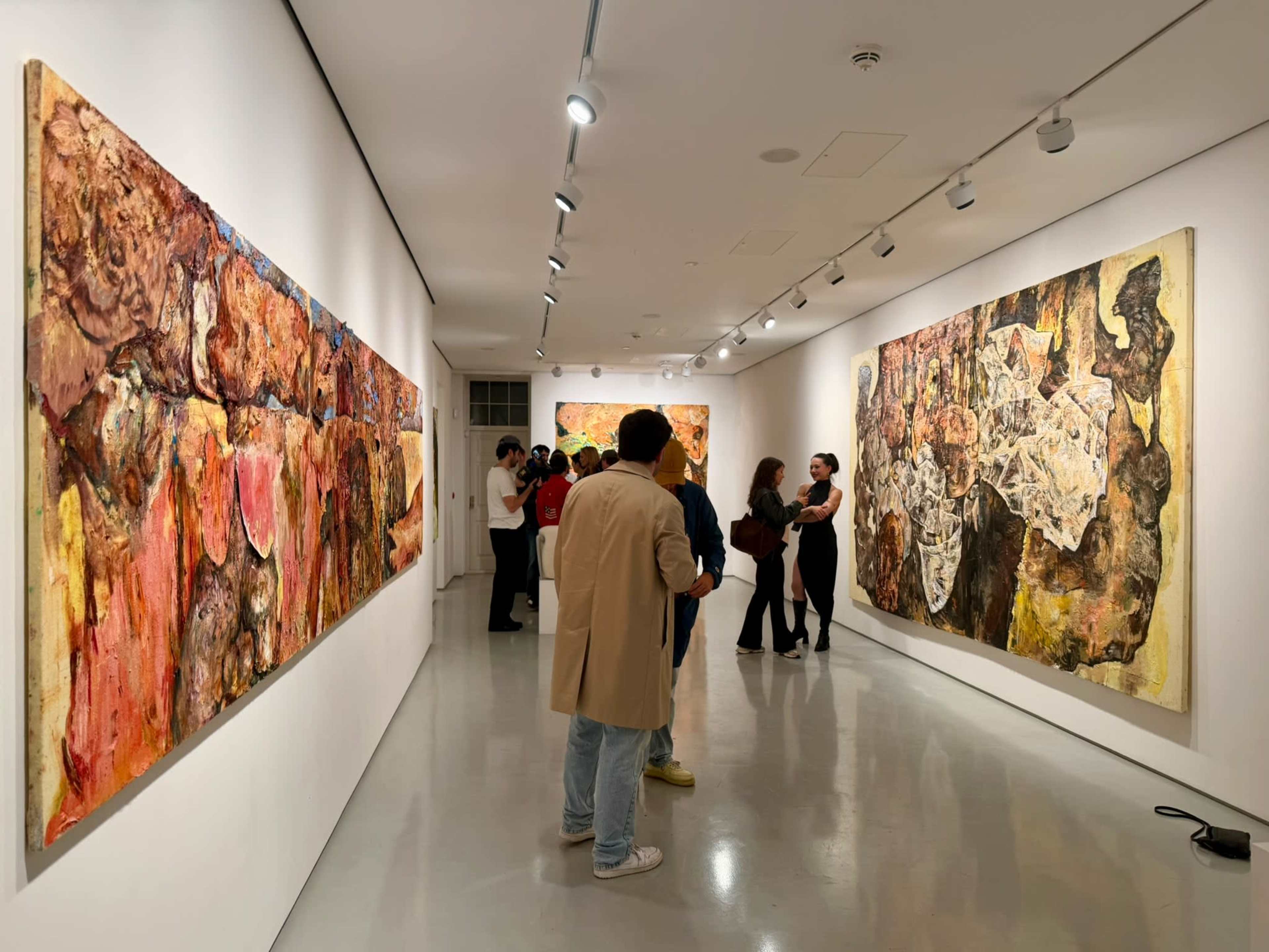 The image shows a gallery space with abstract paintings displayed on the walls and a small group of people admiring the artworks.
