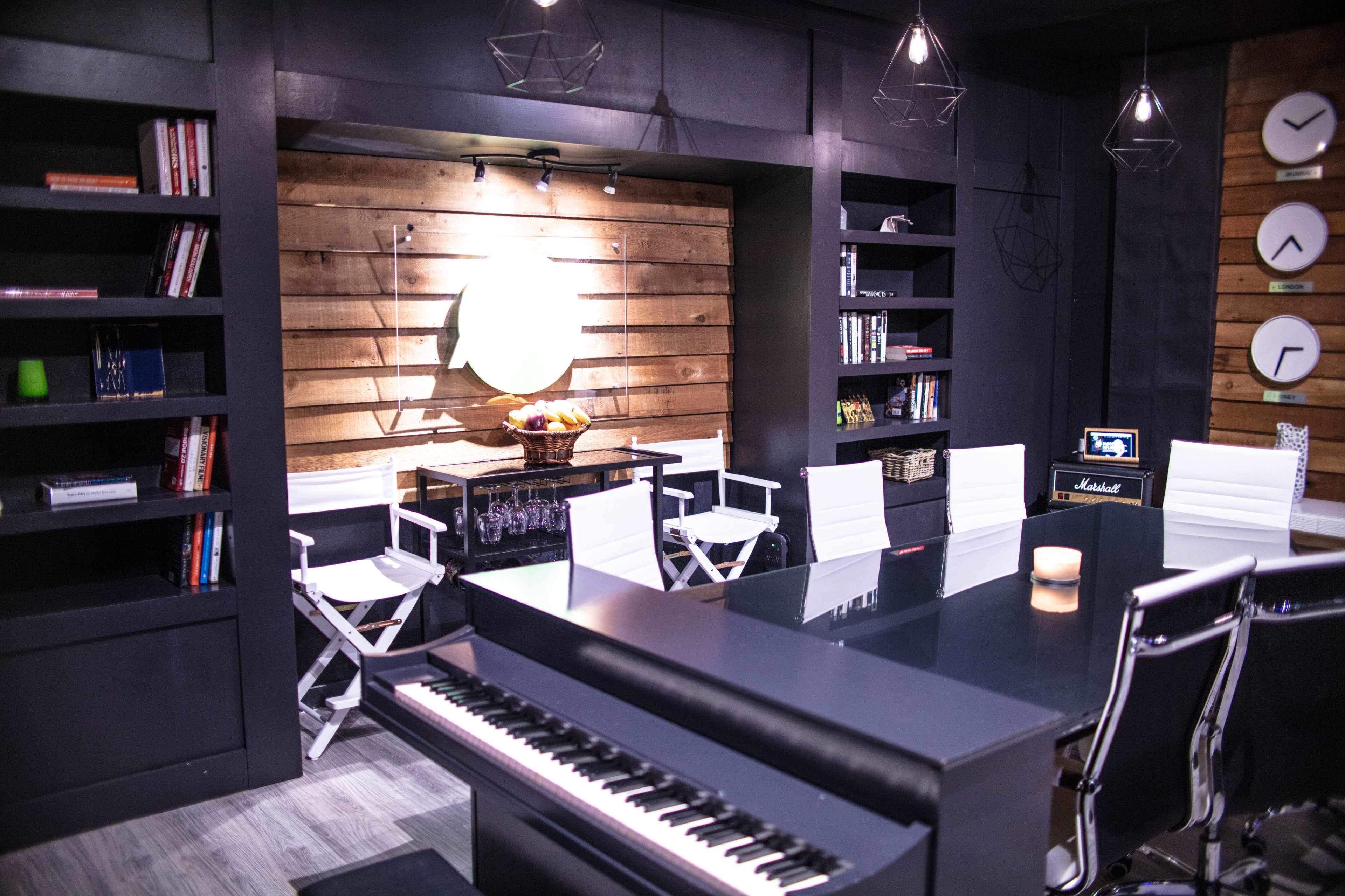 A modern room featuring a piano, a row of white chairs, wooden bookshelves filled with books, and several clocks on the wall.