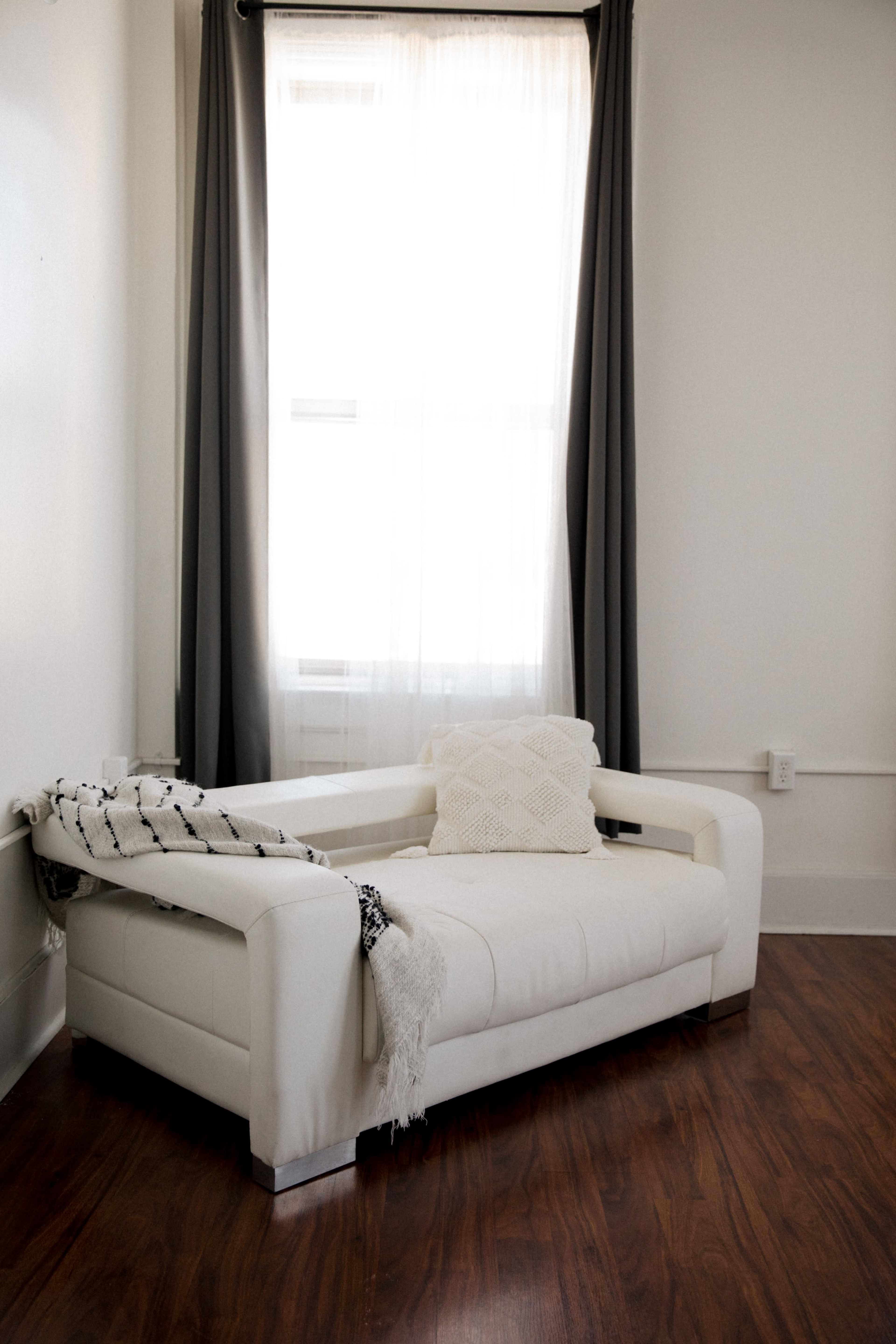 A white sofa with a patterned throw blanket is positioned near a window with sheer curtains in a room with wooden flooring.