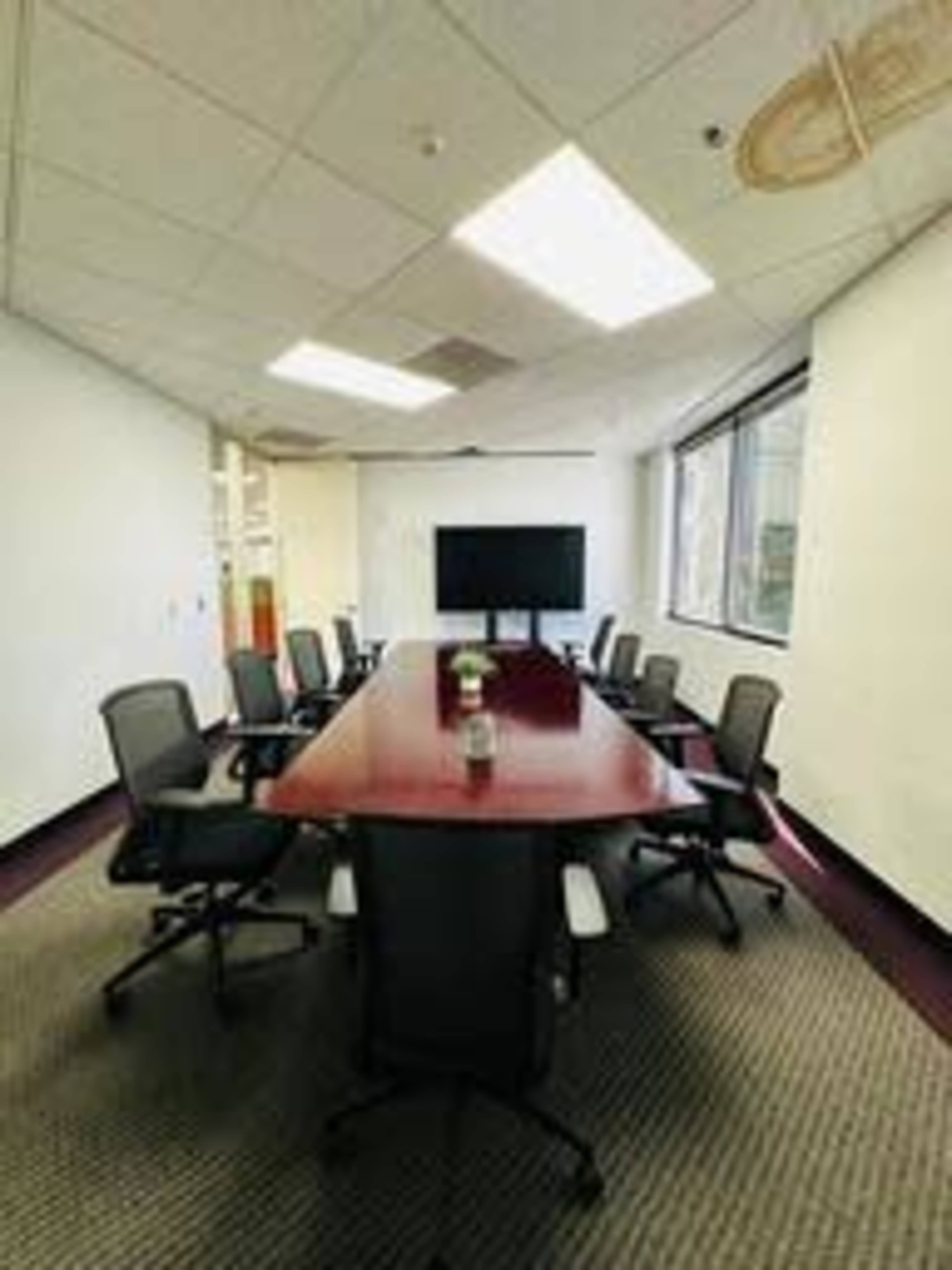 The image shows a conference room with a long, wooden table surrounded by black office chairs and a large screen on one wall.