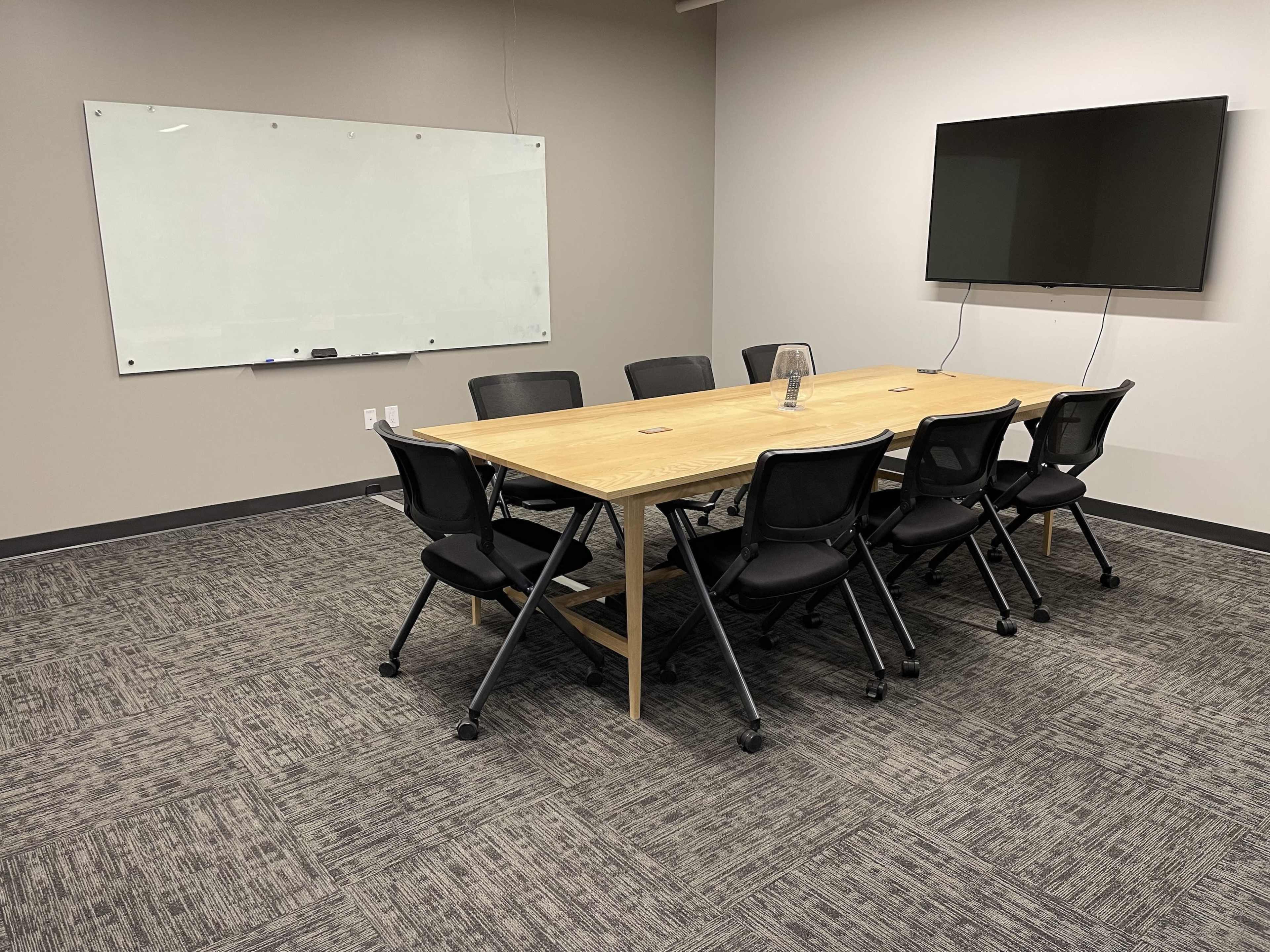 A conference room featuring a long wooden table surrounded by eight black chairs, a whiteboard on the wall, and a mounted television.
