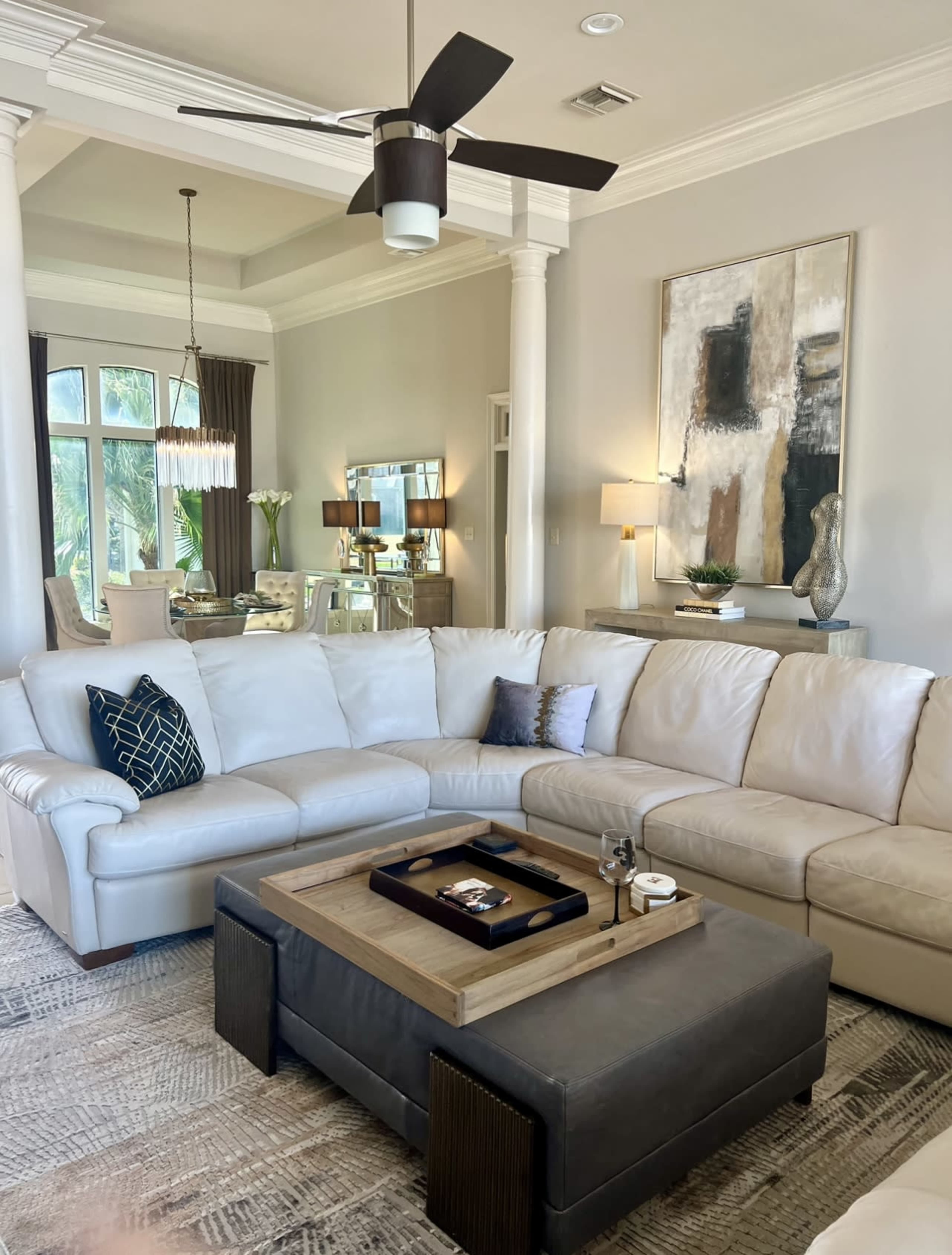 A modern living room featuring a large white sectional sofa, a gray ottoman with a wooden tray, and a dining area visible in the background.