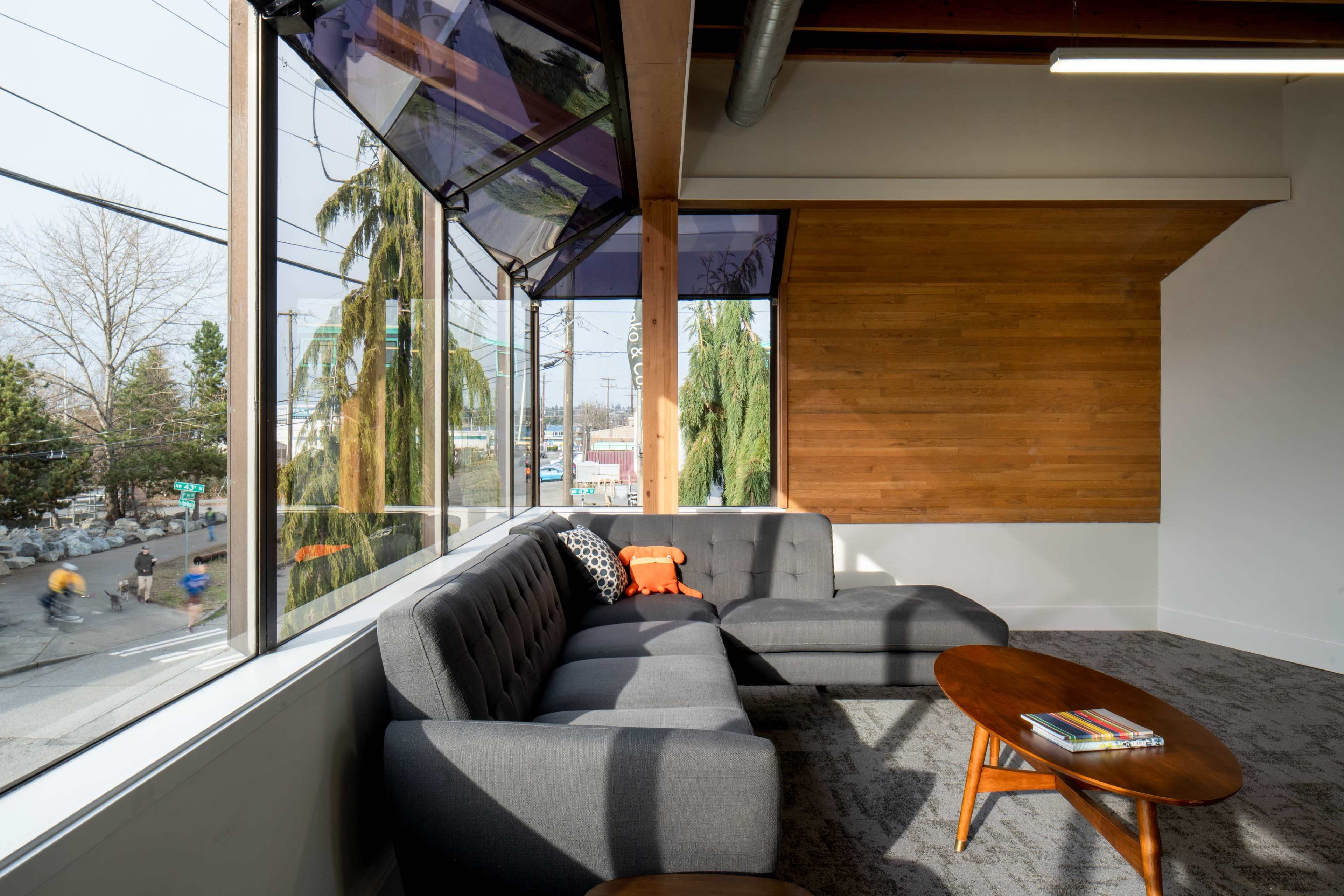 A gray sectional sofa is positioned near large windows, showcasing a view of the street and greenery outside.