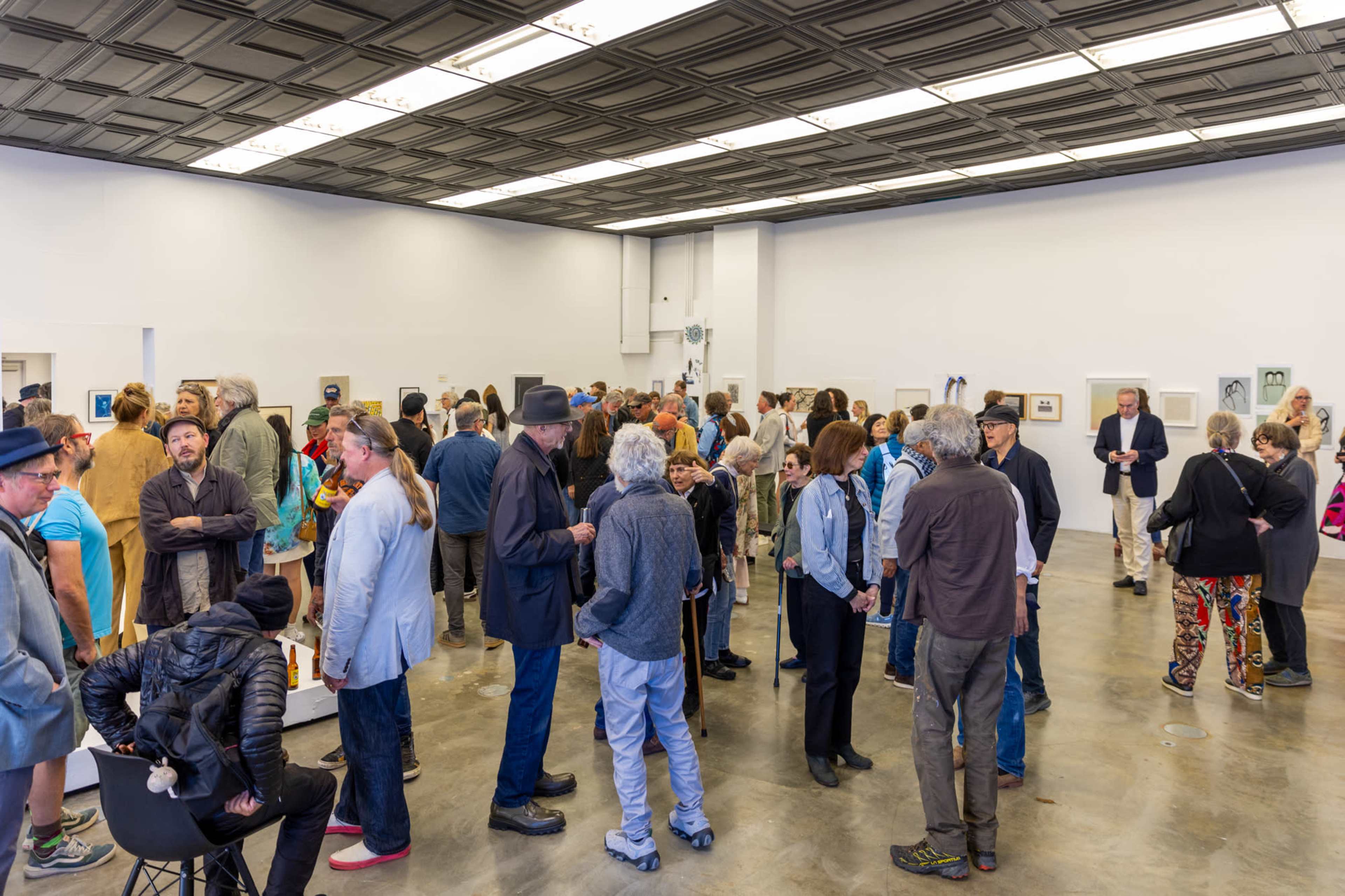 A crowded art gallery features a diverse group of people mingling and viewing the displayed artwork.