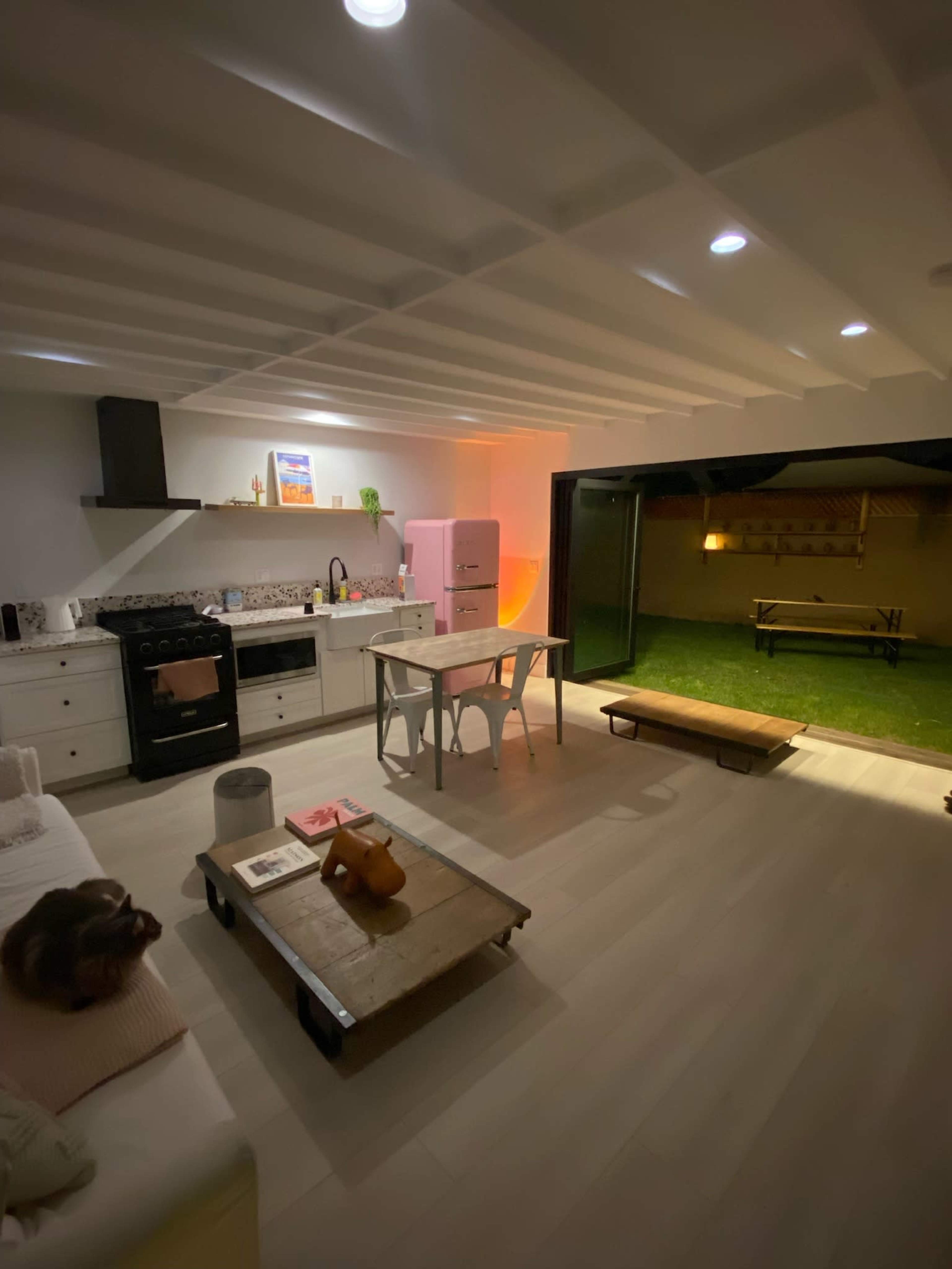 The image shows a modern kitchen and living area with a table and seating, featuring a pink refrigerator and a view of a grassy outdoor space.