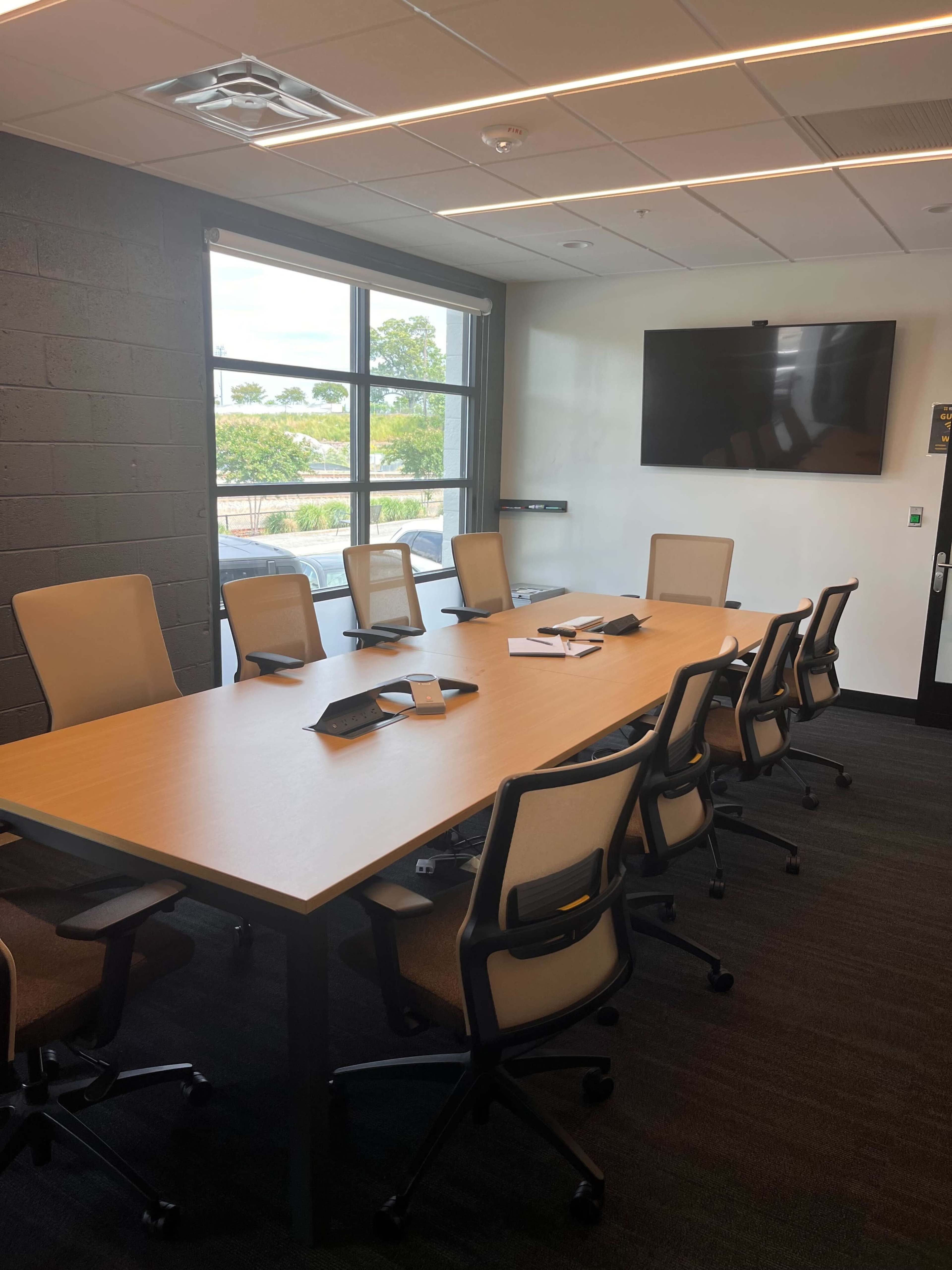 A conference room features a long wooden table surrounded by eight ergonomic chairs, with large windows offering natural light and a wall-mounted screen.